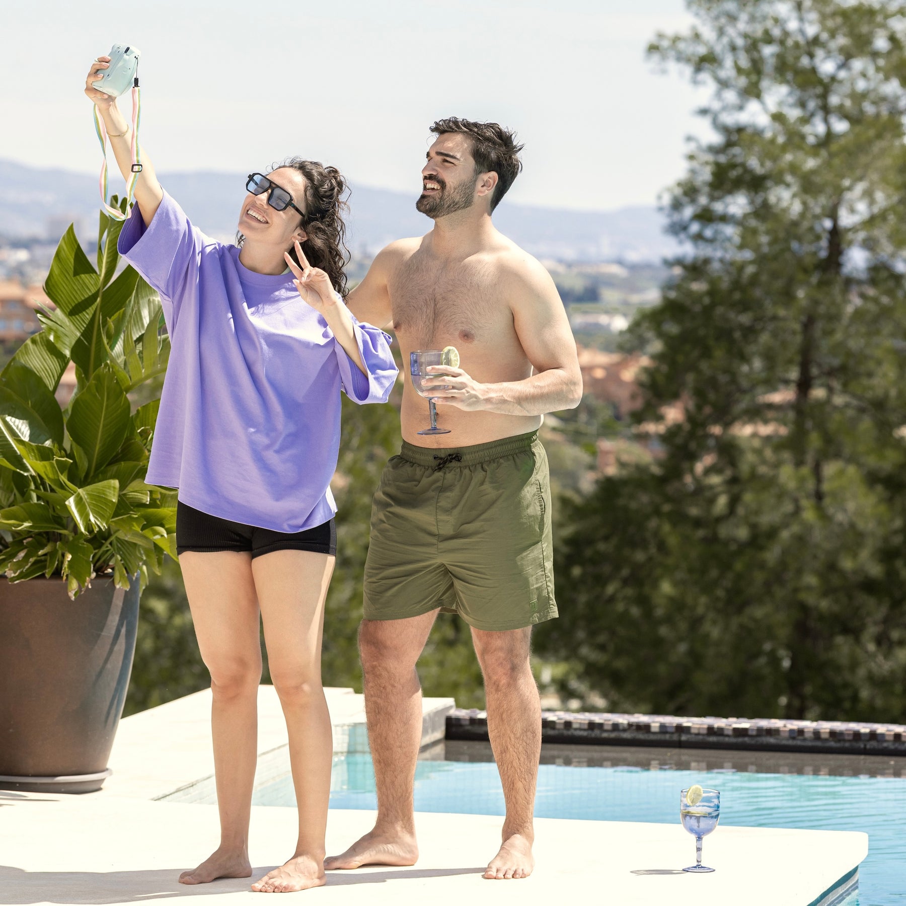 Mann in olivgrüner Badehose 'Jay' steht am Pool neben Frau in lila Oberteil und schwarzen Shorts, die Selfie macht