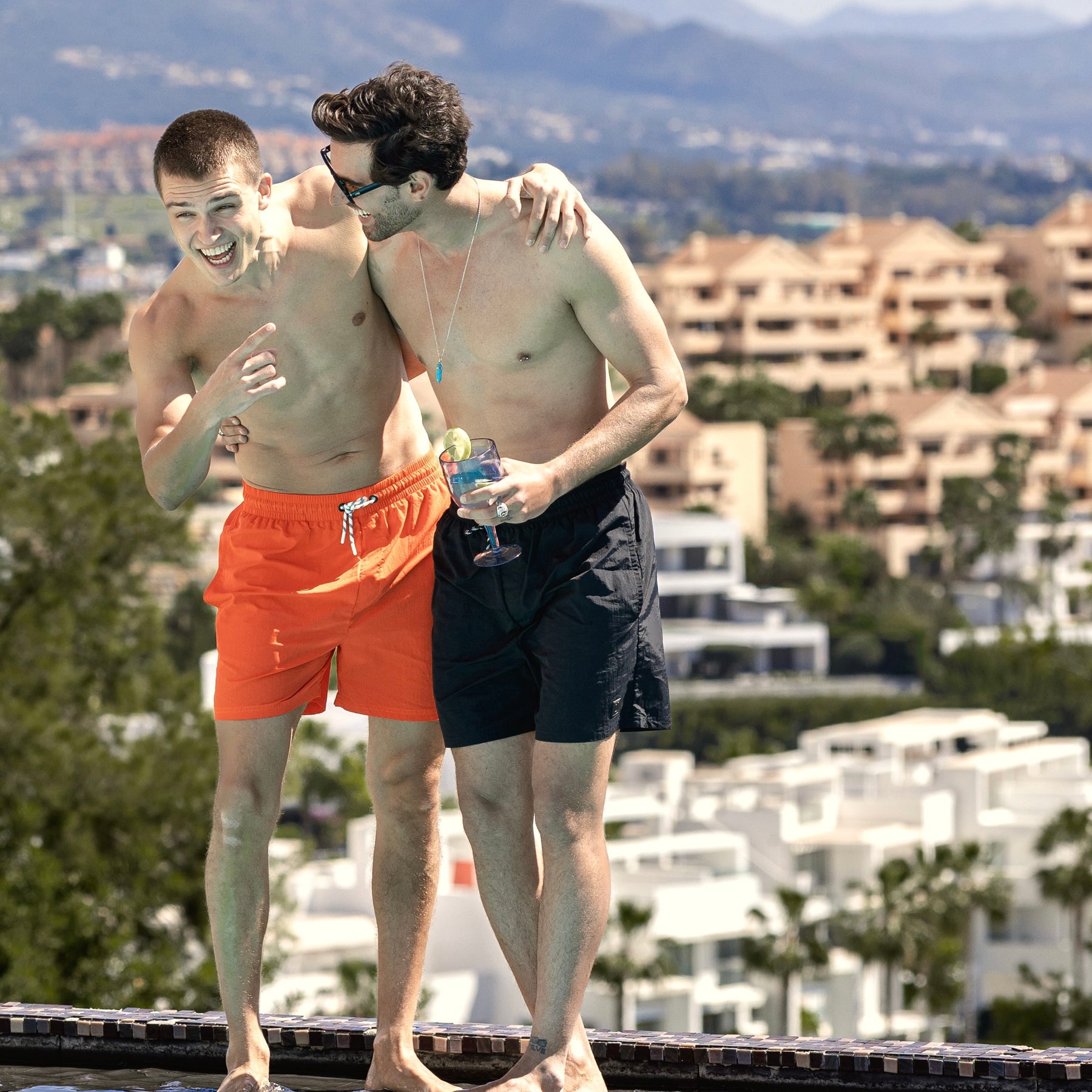 Zwei junge Männer am Pool, einer trägt die schwarze Badehose 'Jay', der andere eine orangefarbene Badehose.