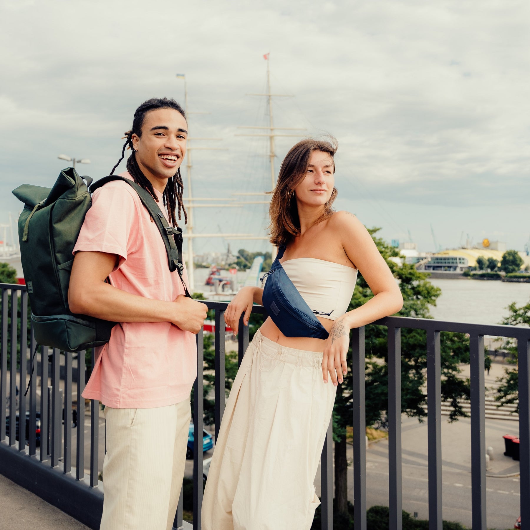 Junge Frau trägt die blaue Bauchtasche 'Erik Small' von Johnny Urban über dem Oberkörper, neben ihr steht ein junger Mann mit grünem Rucksack in urbaner Umgebung