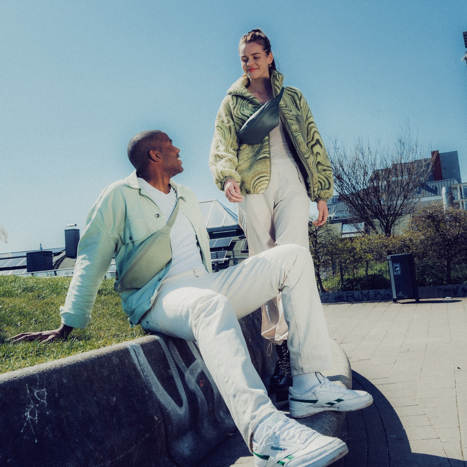 Zwei junge Menschen, ein Mann sitzt auf einer Mauer mit einer hellgrünen Bauchtasche 'Toni' aus Kunstleder, eine Frau steht mit einer schwarzen Bauchtasche 'Toni' aus Kunstleder über der Schulter