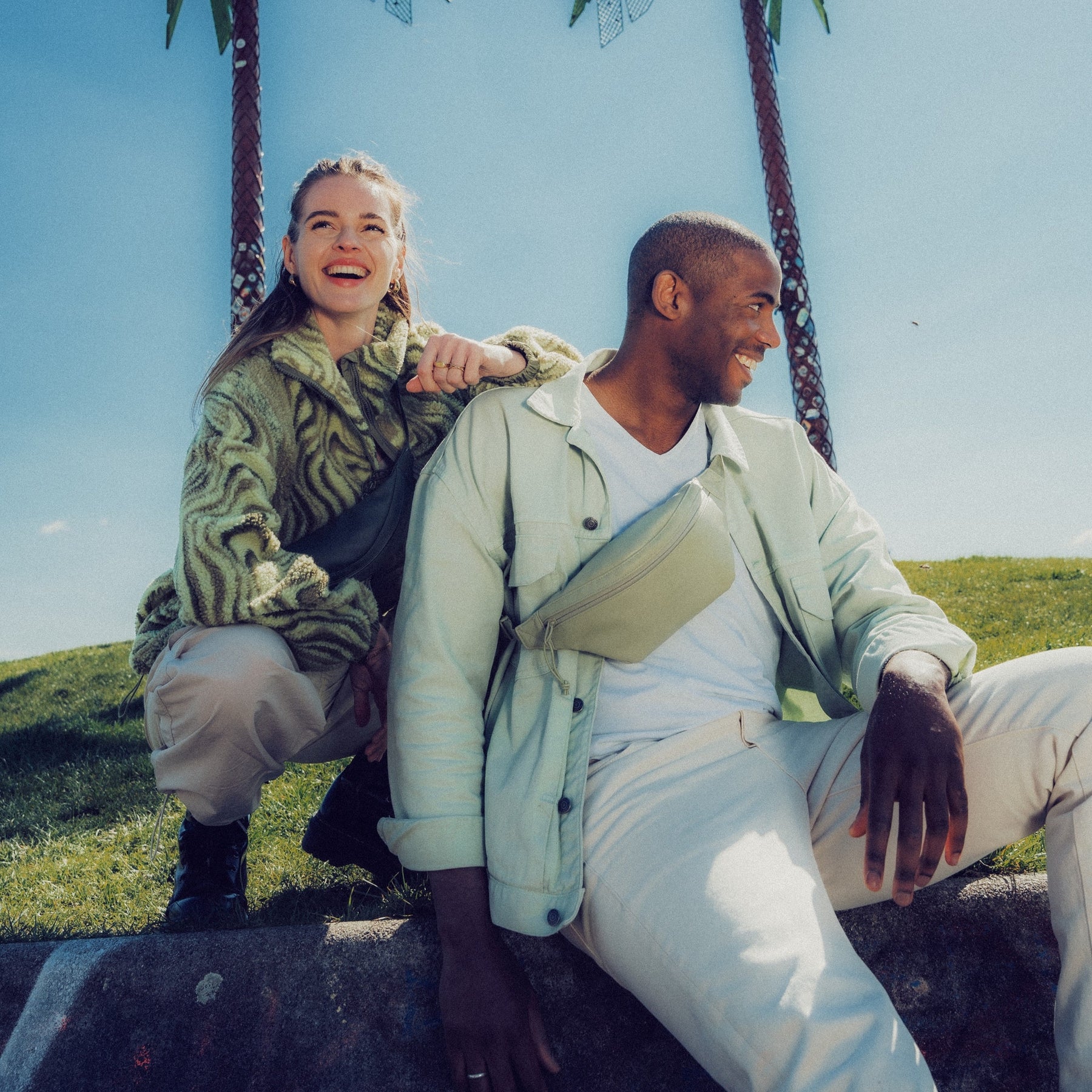 Zwei lachende Personen sitzen draußen, tragen helle Kleidung und je eine limefarbene Bauchtasche 'Toni' aus Kunstleder von Johnny Urban