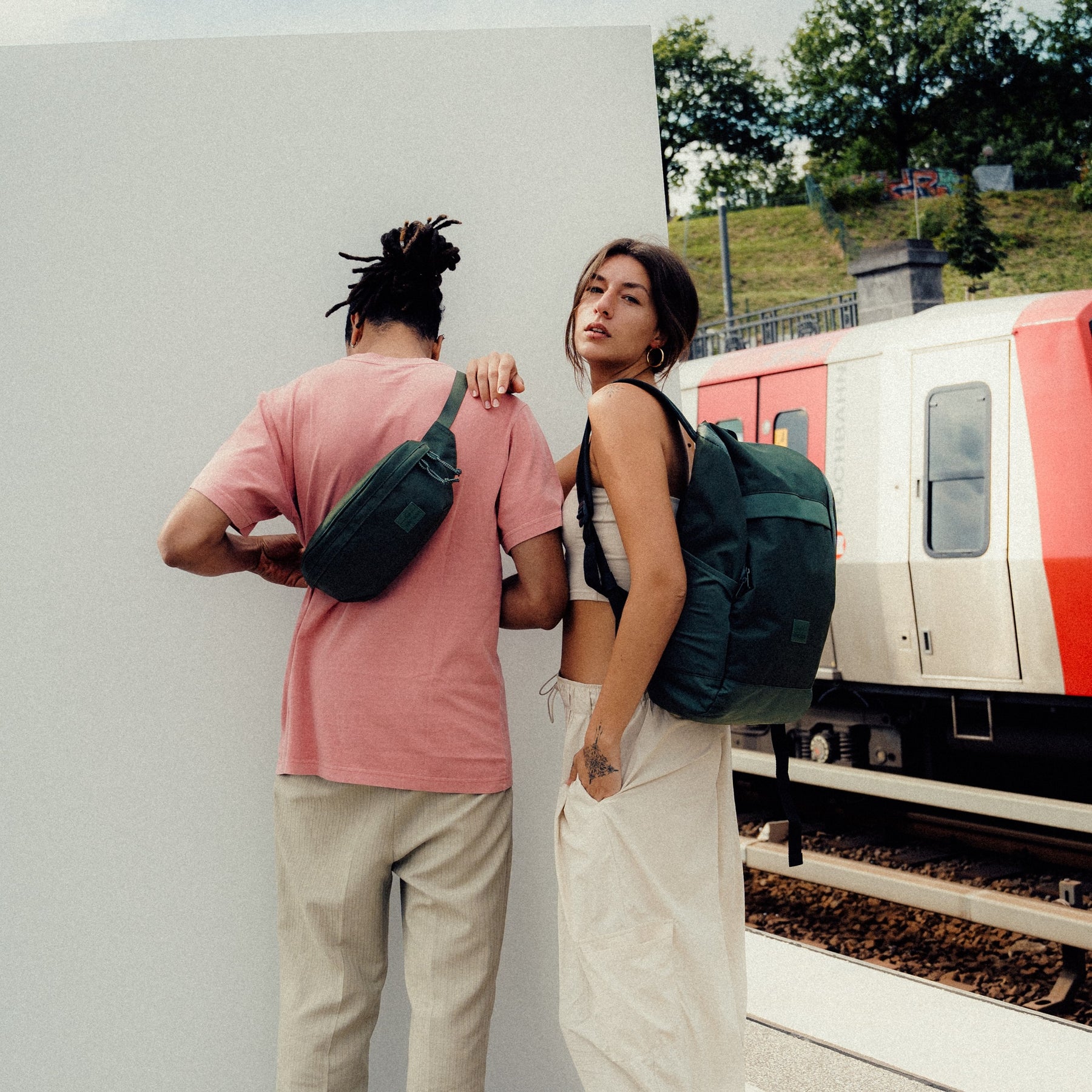 Mann trägt grüne Johnny Urban Bauchtasche 'Tom' quer über dem Rücken mit rosa T-Shirt und Frau mit grünem Rucksack neben Zug