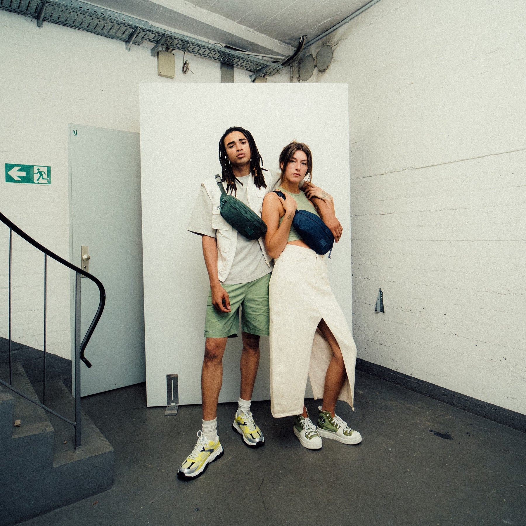 Zwei Personen tragen Johnny Urban Bauchtaschen 'Tom' in Grün und Blau vor weißem Hintergrund in urbanem Innenraum