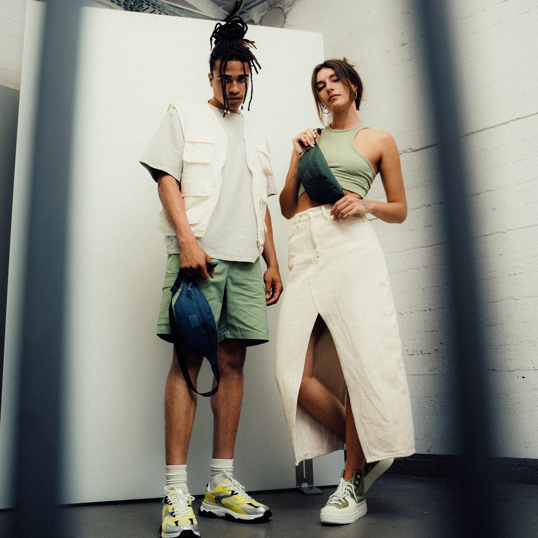 Junger Mann mit grüner Johnny Urban Bauchtasche 'Erik Small' in der Hand, junge Frau trägt gleiche Bauchtasche schräg über die Schulter, beide urban gekleidet