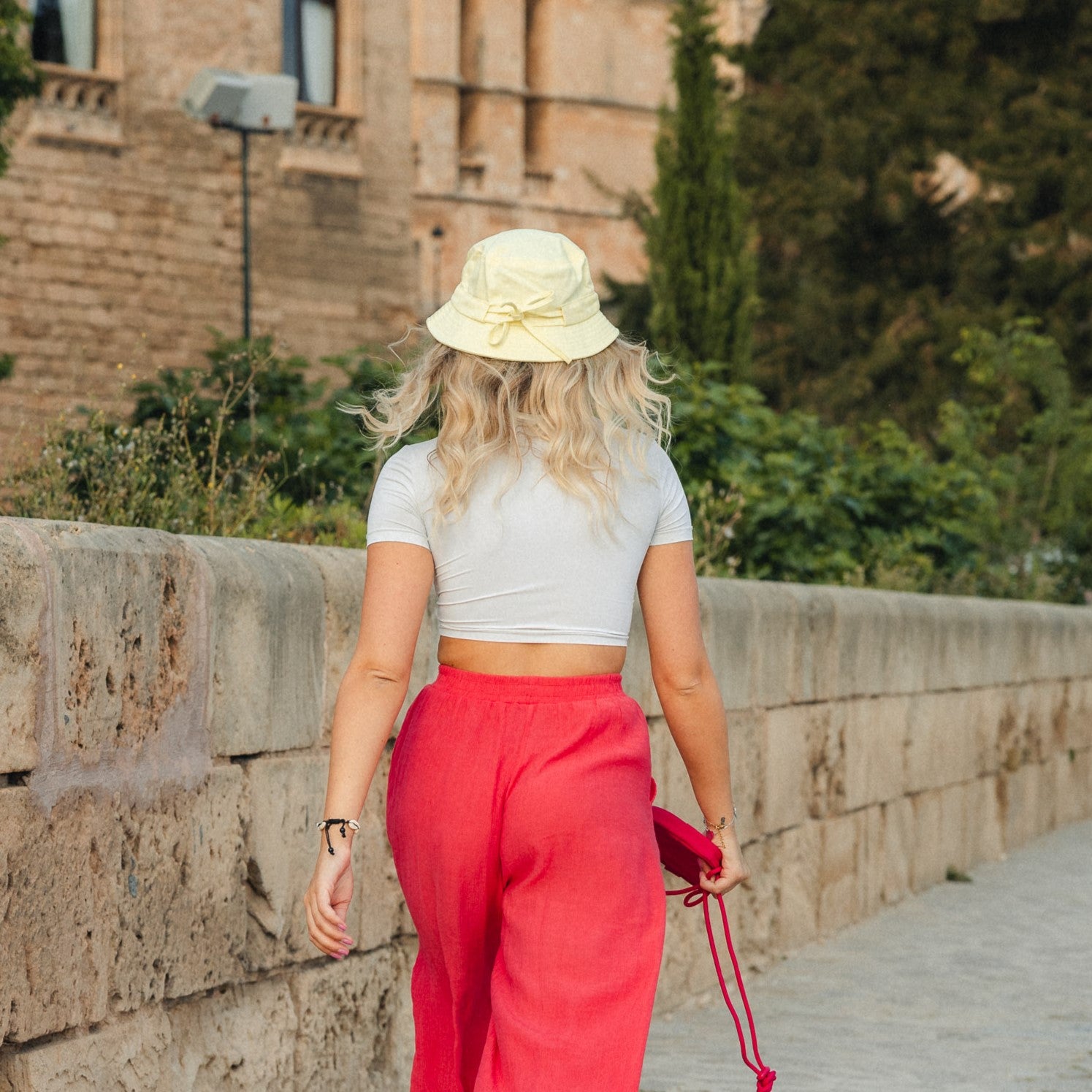 Frau von hinten mit gelbem Bucket Hat 'Gill', weißem Crop-Top und roten Hosen, die an einer Steinmauer vorbeigeht