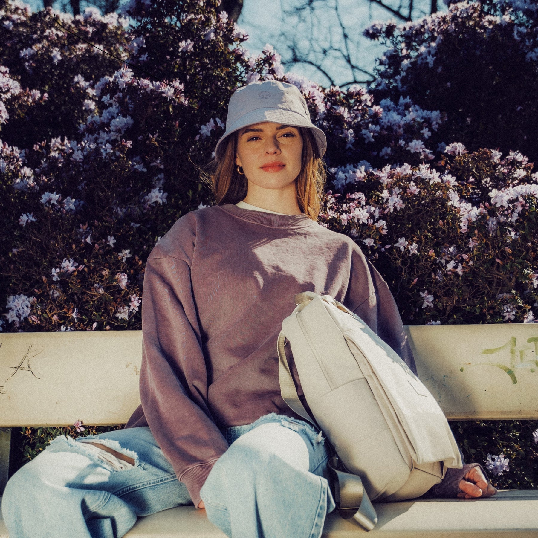 Frau mit lila Sweatshirt, hellbeiger Bucket Hat 'Bob' und cremefarbenem Rucksack sitzt auf Parkbank vor blühendem Strauch