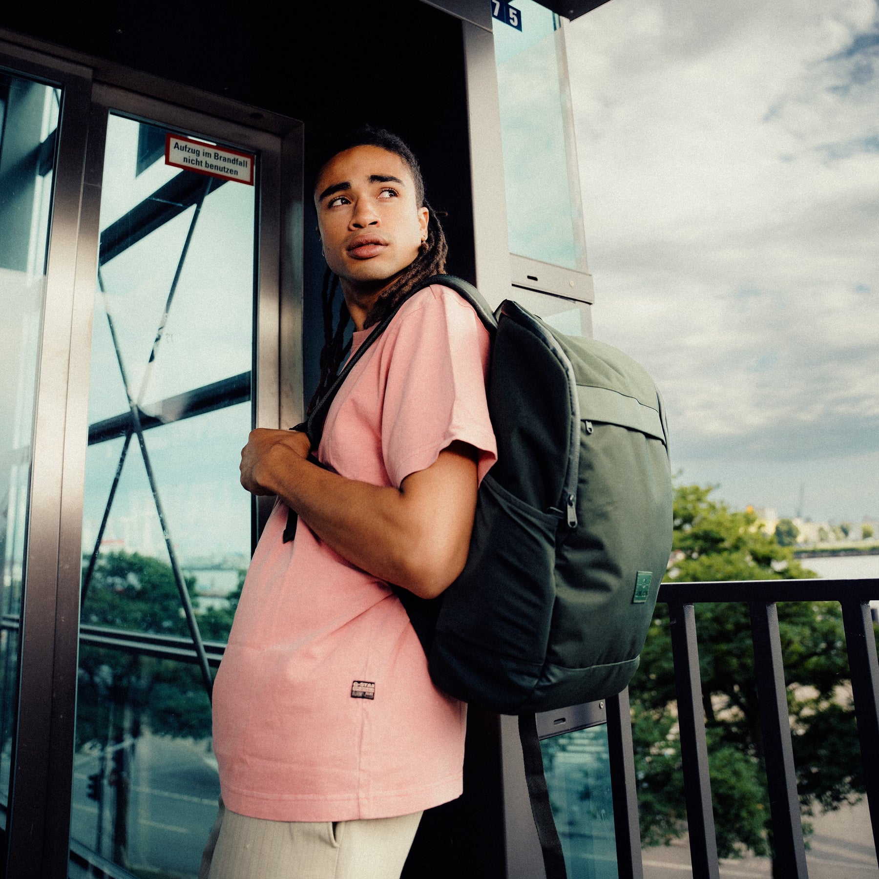 Mann mit Business-Rucksack 'Jasper' von Johnny Urban in Olivgrün und rosa T-Shirt stehend an einer Glasscheibe im urbanen Umfeld