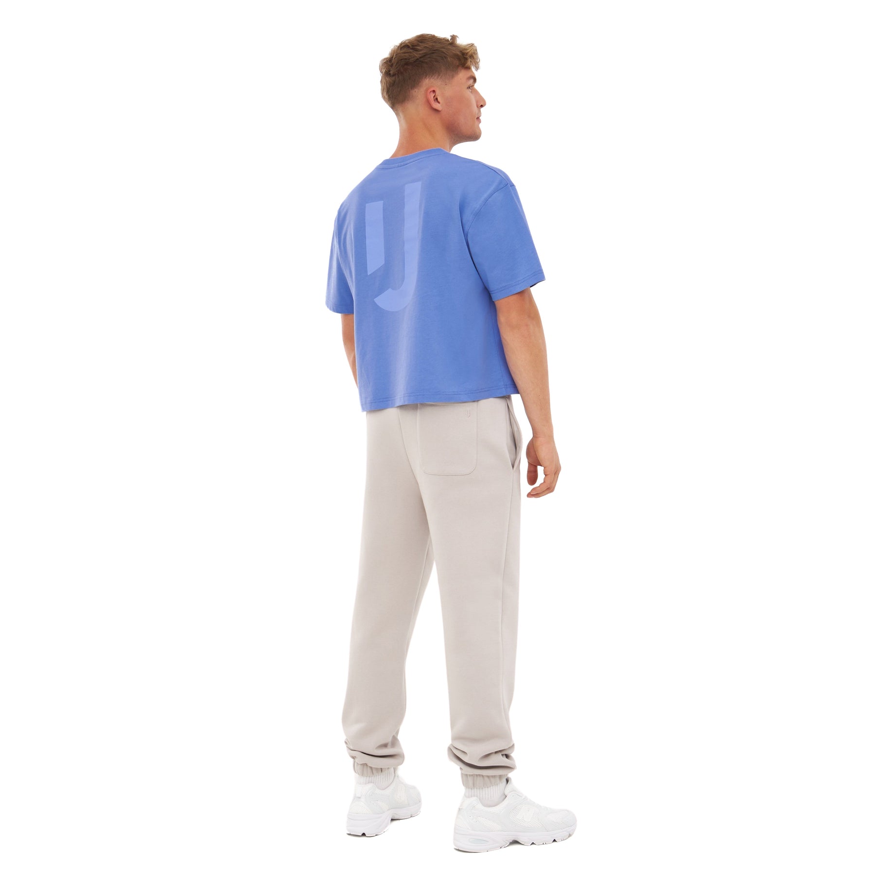Mann trägt blaues, oversize Cropped T-Shirt 'Zoey JU' mit kurzem Ärmel und Logo auf dem Rücken, beige Jogginghose und weiße Sneaker