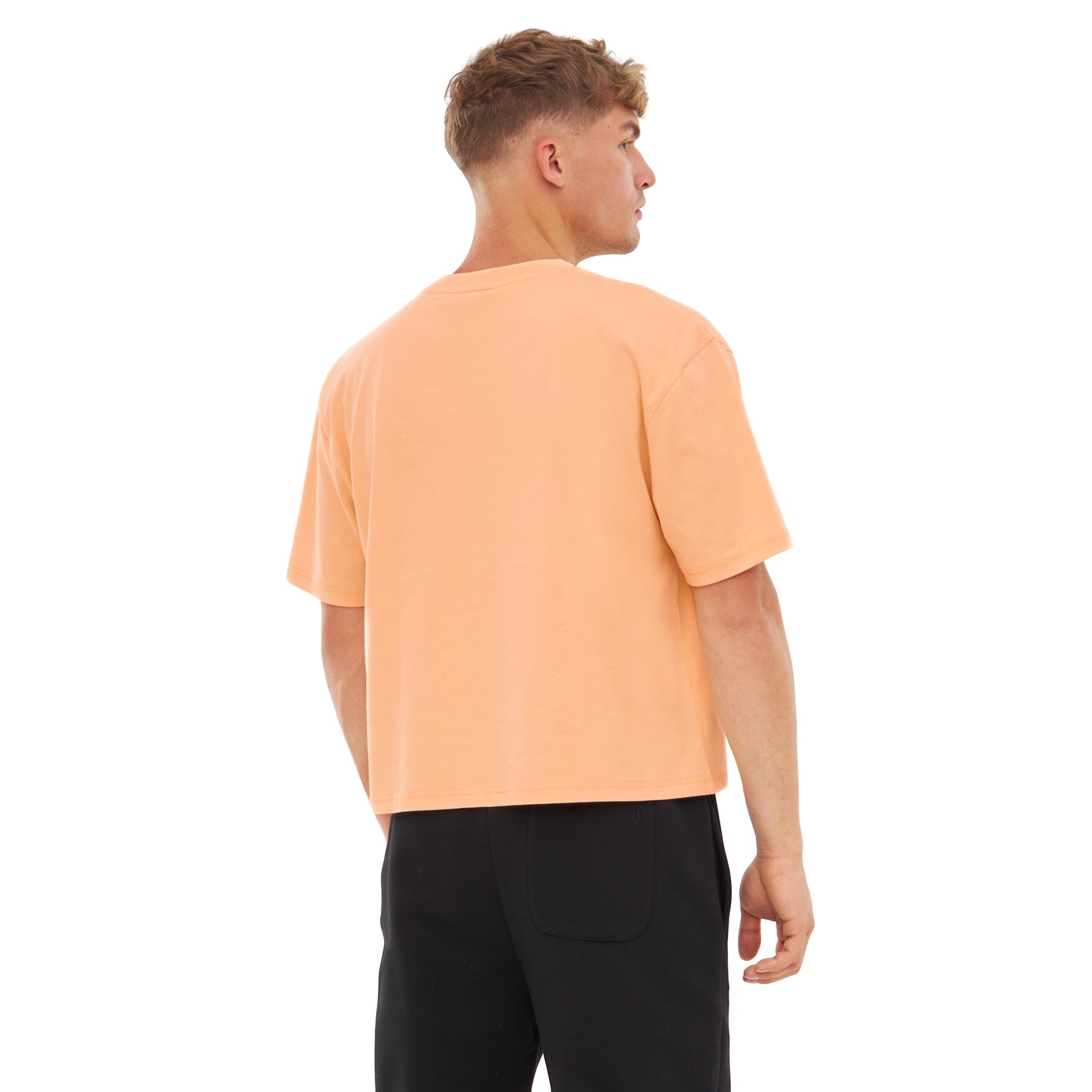 Mann trägt 'Zoey' Oversize Cropped T-Shirt in Pfirsich, Rückansicht, kombiniert mit schwarzen Hosen
