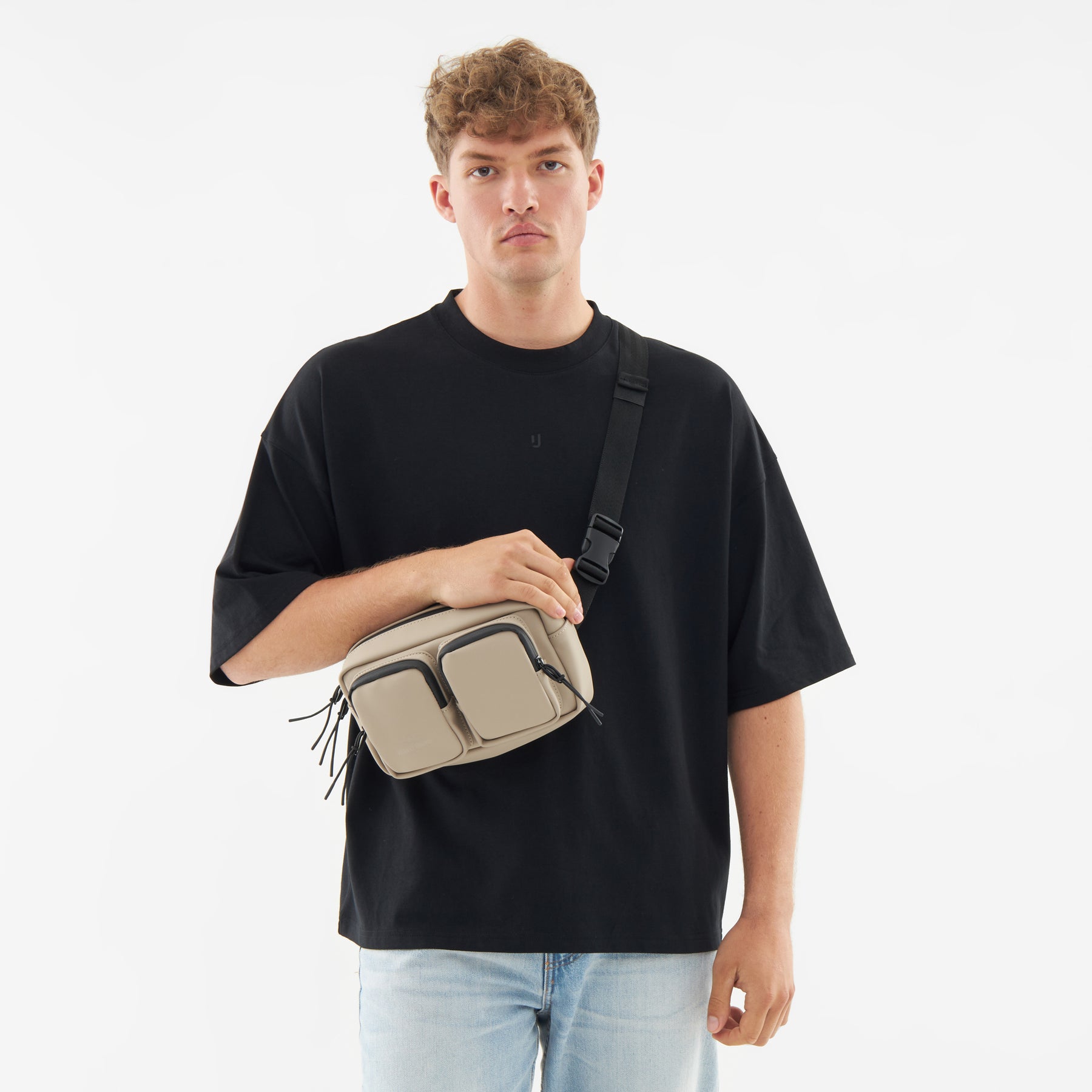 Beige Umhängetasche 'Wesley' aus Stoff mit mehreren Reißverschlusstaschen, getragen von einem Mann in schwarzem T-Shirt und blauer Jeans