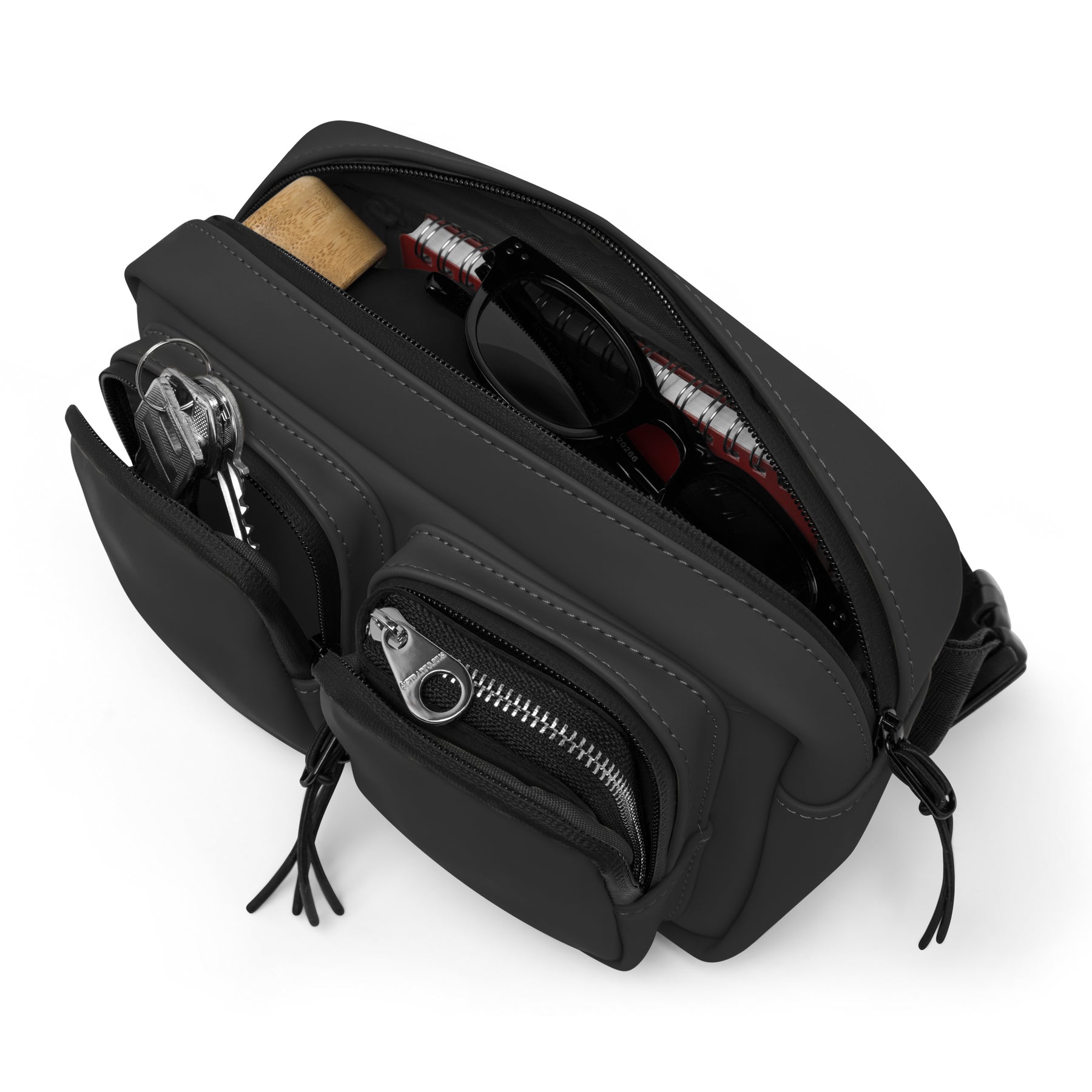 Schwarze Umhängetasche ‚Wesley‘ mit Reißverschlusstaschen und sichtbaren Inhalten Schlüssel, Sonnenbrille, Notizbuch, Lippenstift