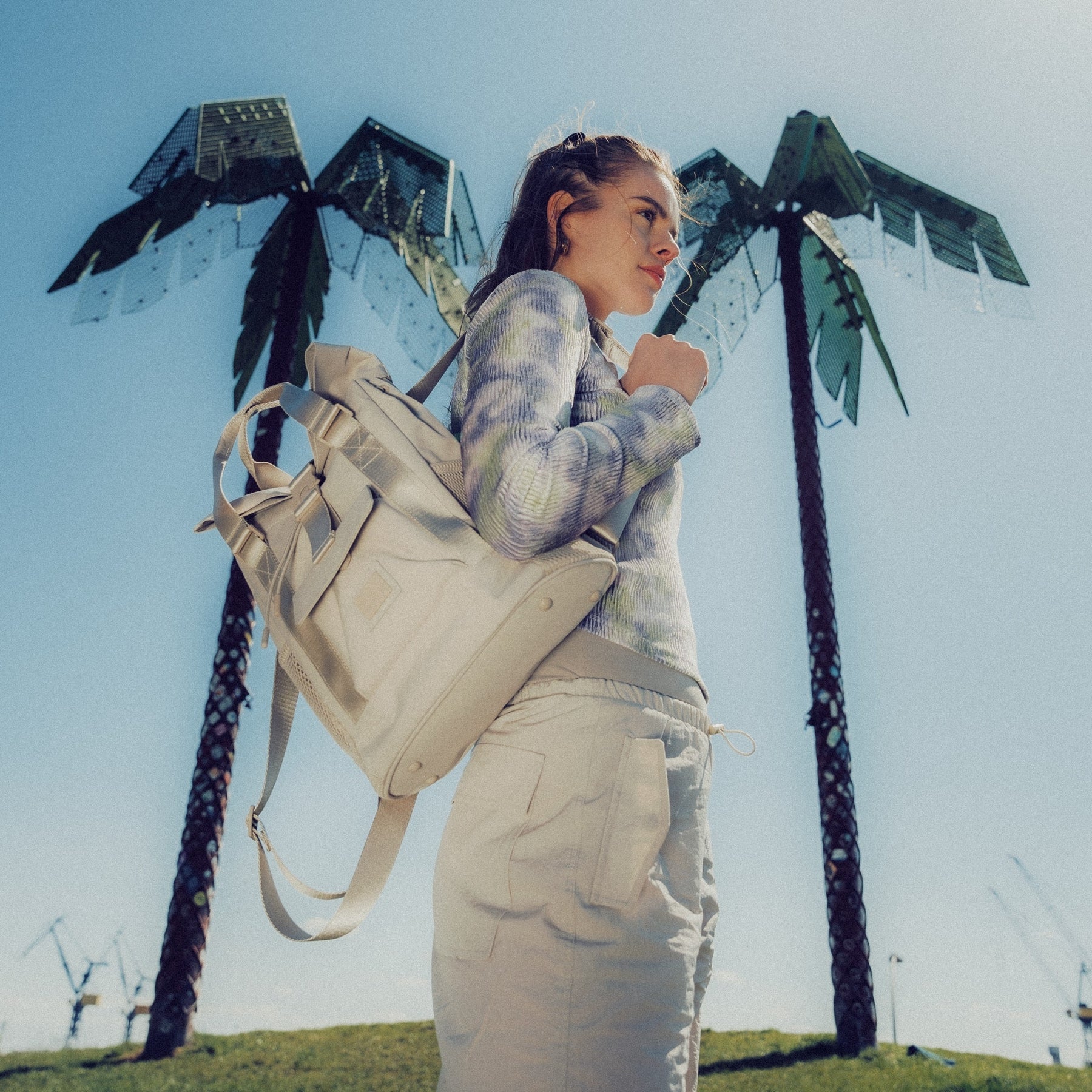 Frau mit beigeem Rucksack 'Jules' und beiger Hose vor blassem Himmel mit zwei hohen Skulpturen