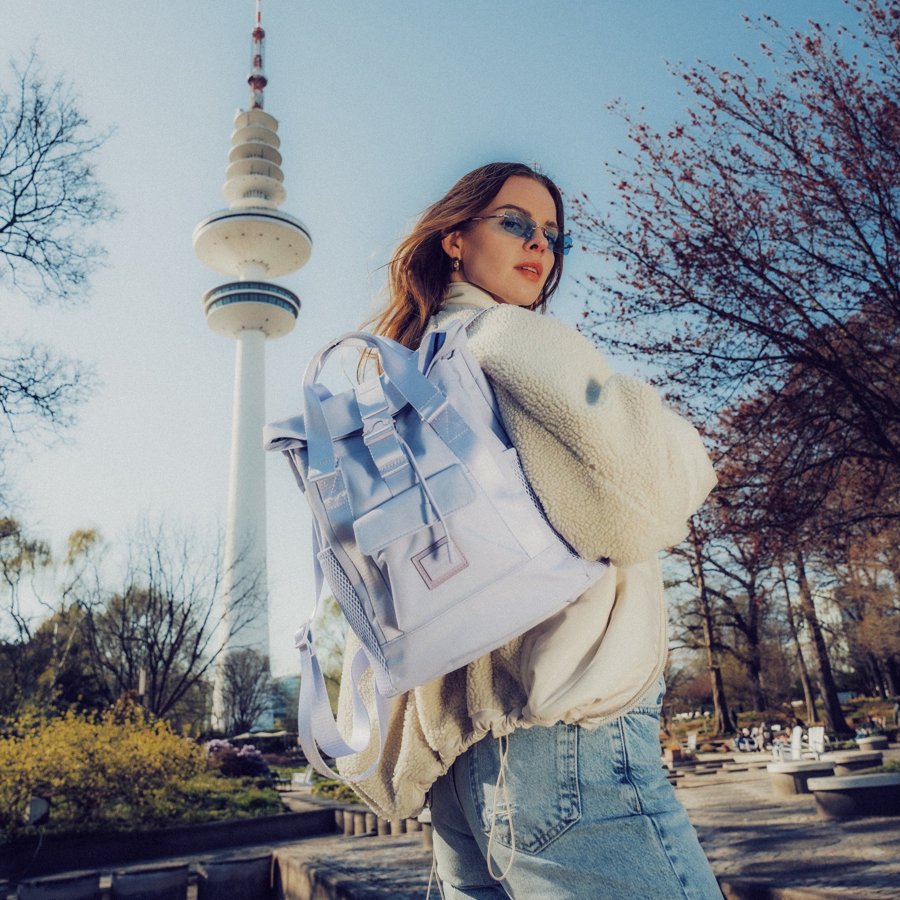 Frau mit lila Damenrucksack 'Jules' trägt weiße Jacke und Jeans vor Fernsehturm im Park