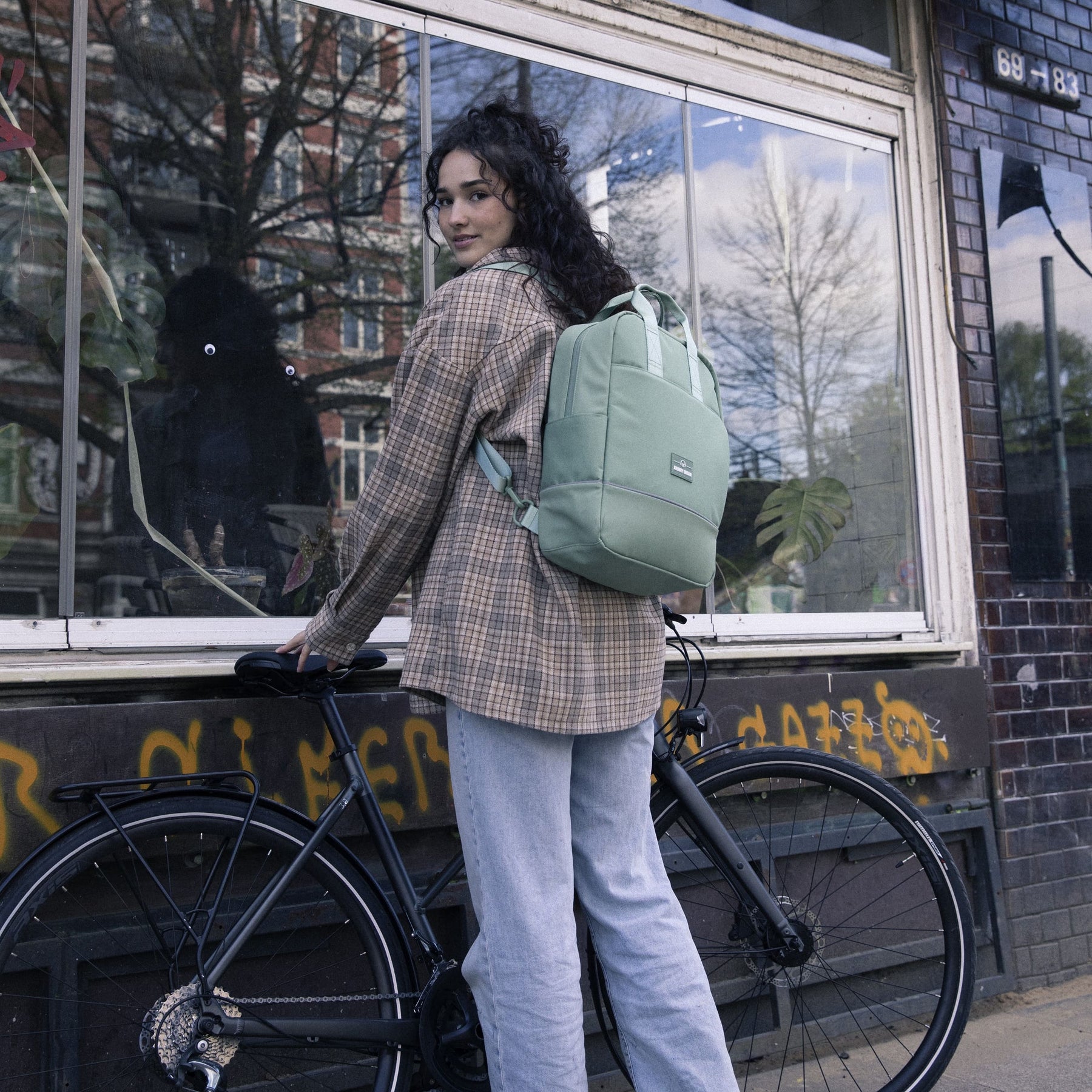 Frau mit mintgrünem Johnny Urban Rucksack Jona Medium sitzt auf Fahrrad vor Fenster mit Pflanzen