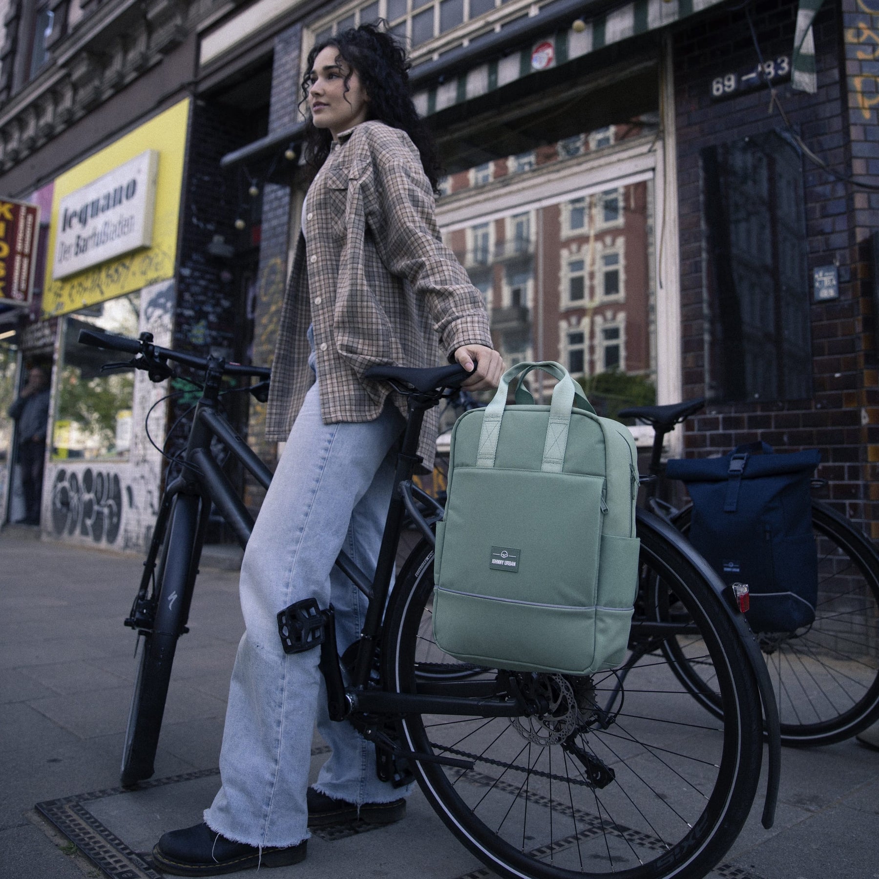 Frau lehnt an schwarzem Fahrrad mit grünen Rucksack 'Jona Medium' von Johnny Urban am Gepäckträger
