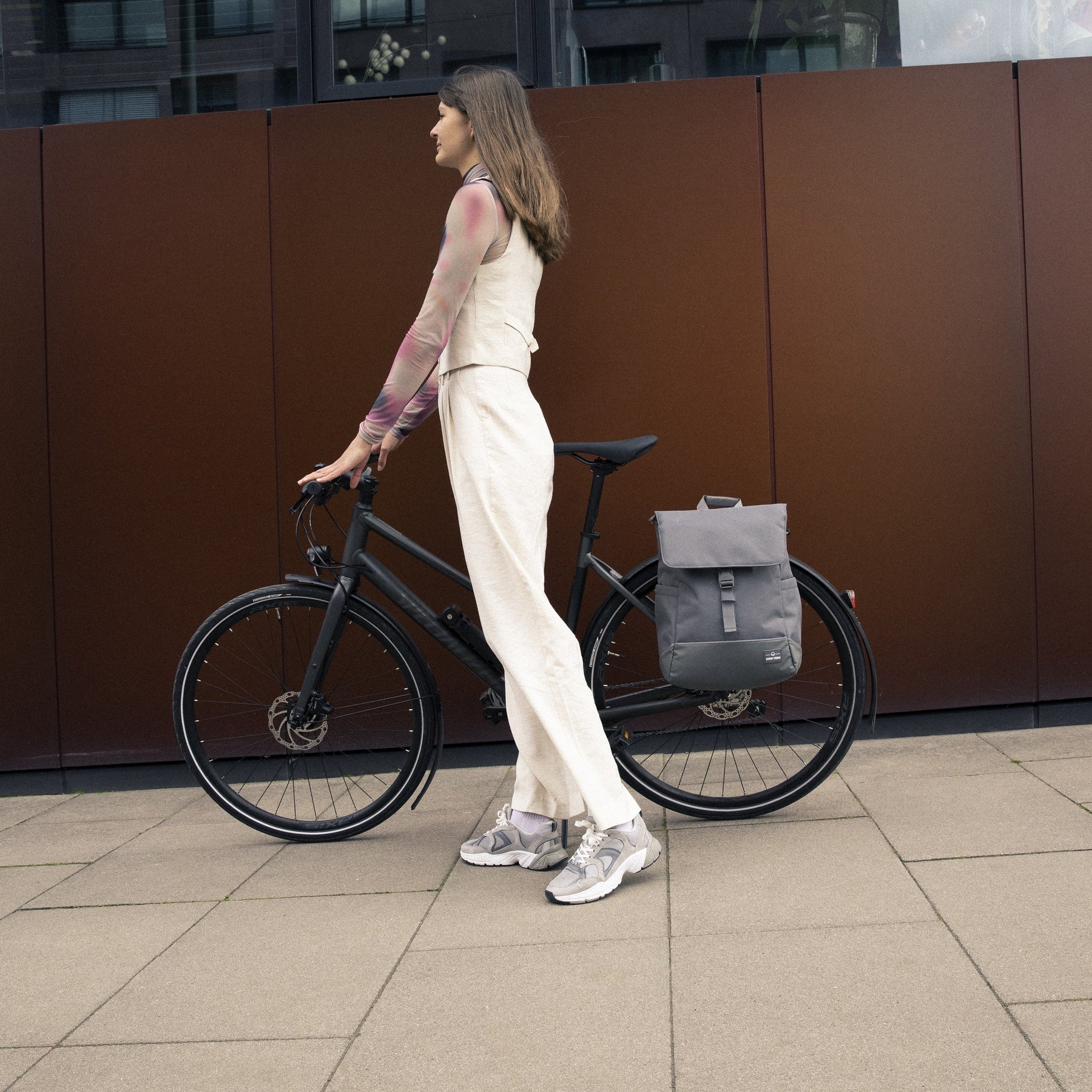 Frau in heller Kleidung steht neben schwarzem Fahrrad mit grauer Fahrradtasche Rucksack 'Mika' Bike am Gepäckträger