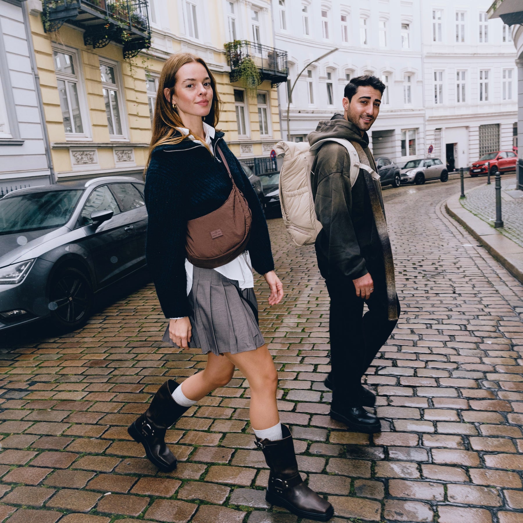 Frau geht auf Kopfsteinpflaster mit brauner Half Moon Bag »Kaia Medium«, Mann trägt Rucksack und Kapuzenpullover
