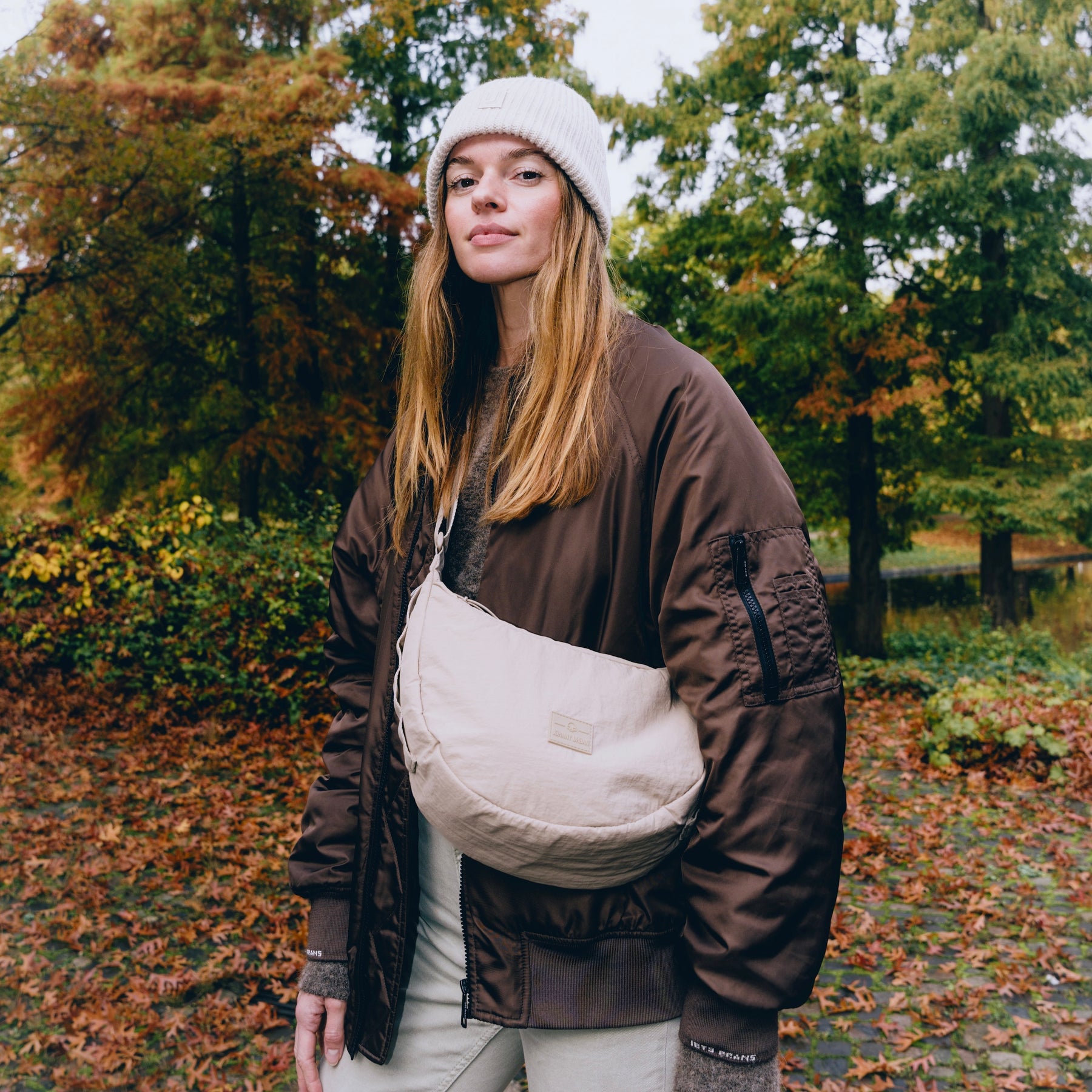 Frau mit beige gepolsterter Half Moon Bag 'Kaia Medium', brauner Jacke und weißer Mütze im herbstlichen Park