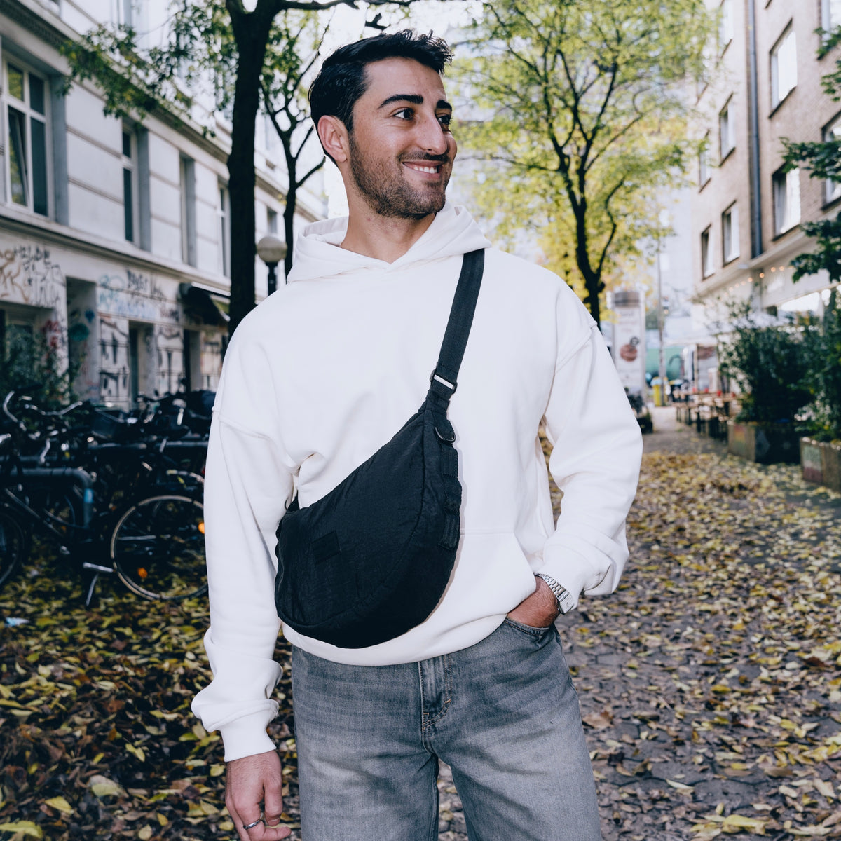 Mann mit schwarzer Half Moon Bag 'Kaia Medium', weißem Hoodie und blauen Jeans auf herbstlicher Straße