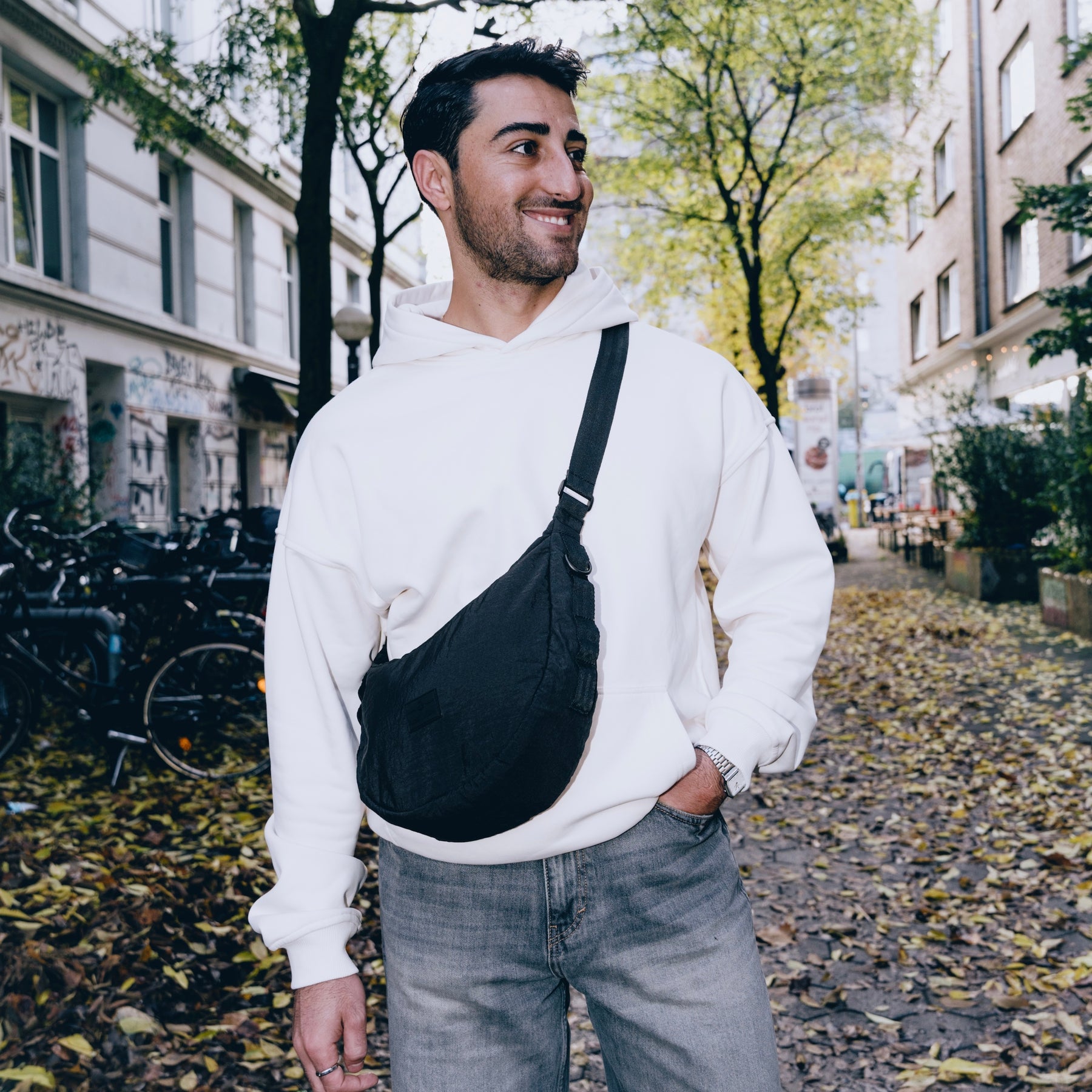 Mann mit schwarzer Half Moon Bag 'Kaia Medium', weißem Hoodie und blauen Jeans auf herbstlicher Straße
