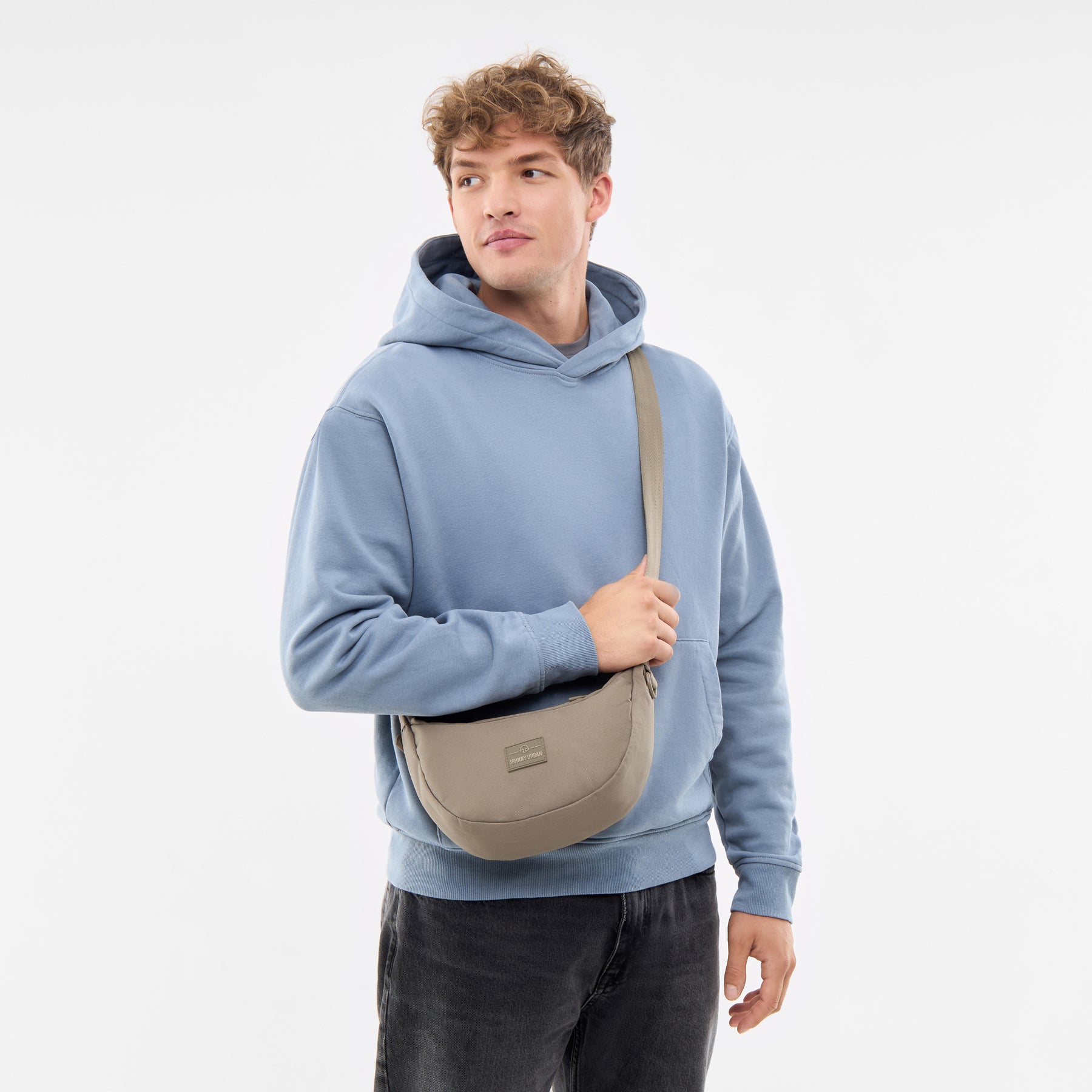 Mann trägt kleine Half Moon Bag 'Ally Small' in desert-grau über der Schulter, blaue Kapuzenjacke, schwarze Jeans
