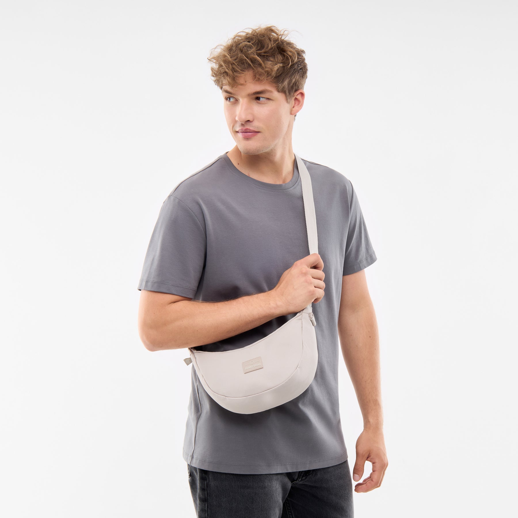 Mann trägt Johnny Urban Half Moon Bag 'Ally Small' in Sandfarben über die Schulter, grau T-Shirt, schaut seitlich