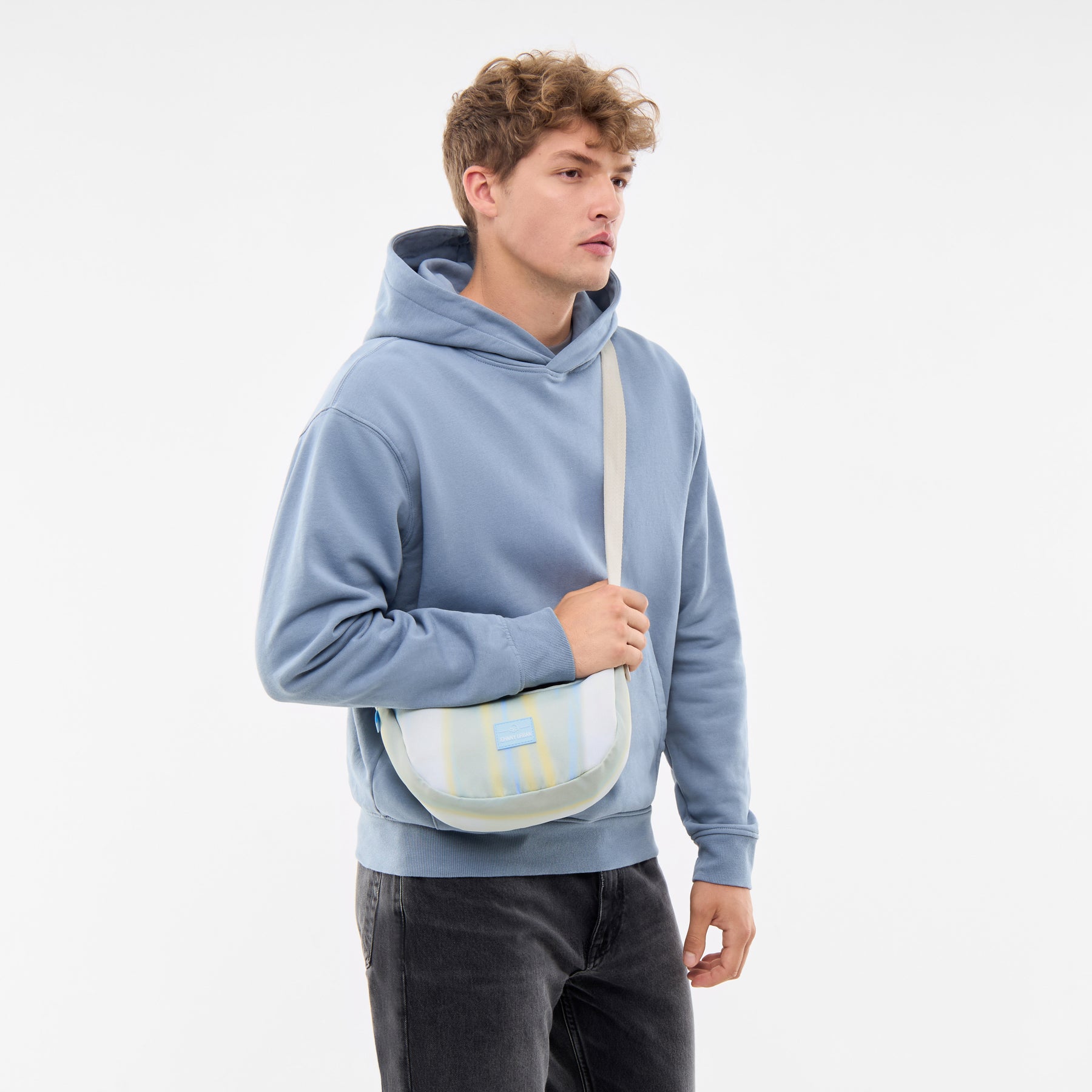 Mann trägt kleine Half Moon Bag 'Ally Small' in Sand mit Streifen und hellblauen Hoodie