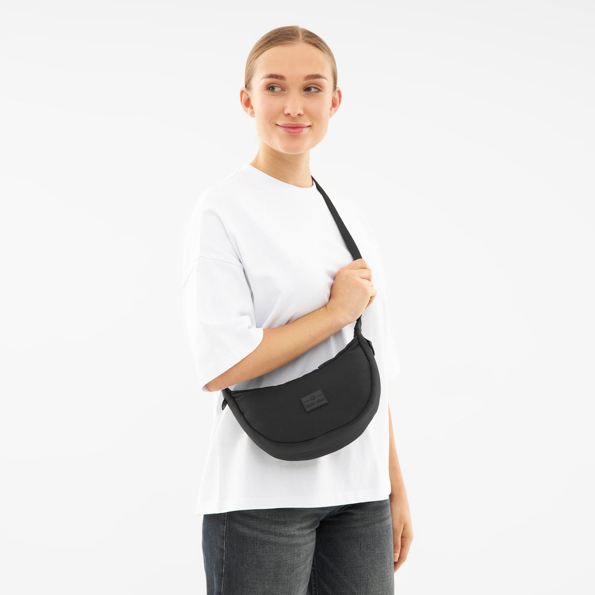 Frau trägt schwarze Half Moon Bag Kaia Small quer am Körper, weißes Oberteil, dunkle Jeans