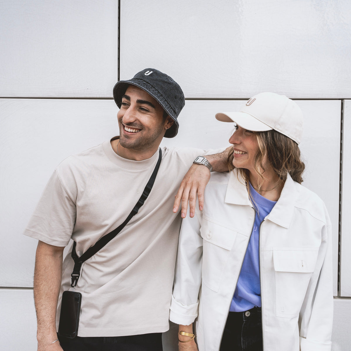 Zwei Personen stehen nebeneinander vor einer weißen Wand, eine trägt eine schwarze Handykette um den Hals