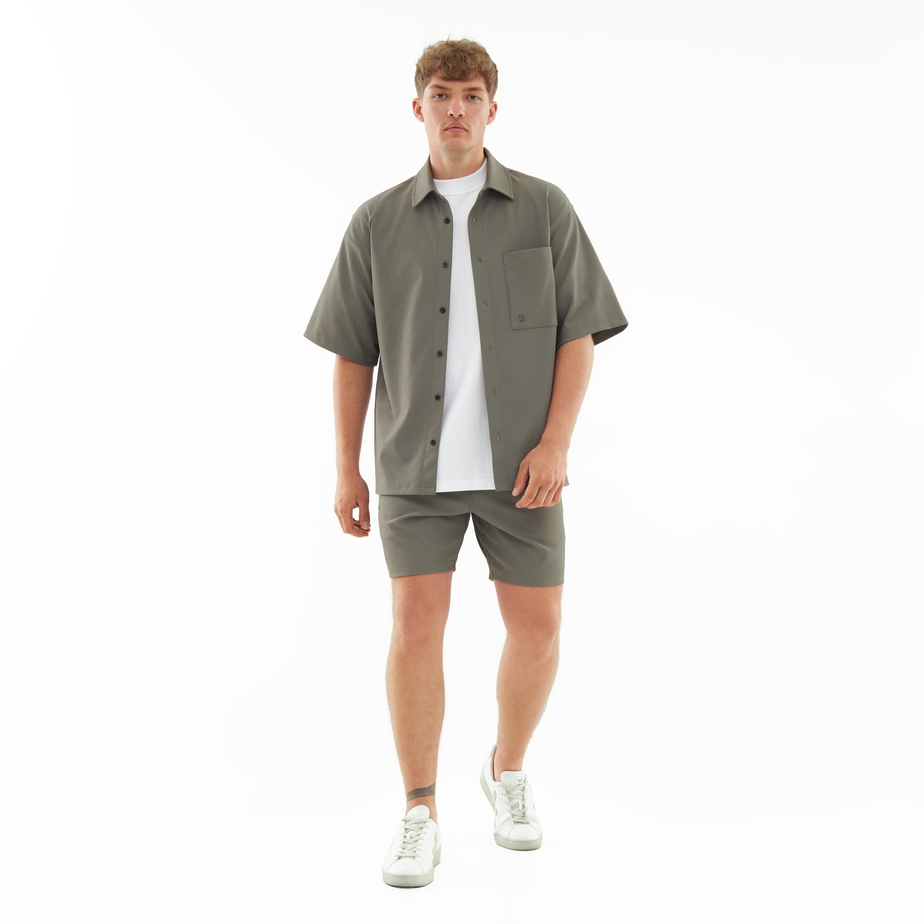 Männliches Model trägt lockeres, olivgrünes Hemd und passende Shorts aus dem Set 'Mason' mit weißem T-Shirt und weißen Sneakern