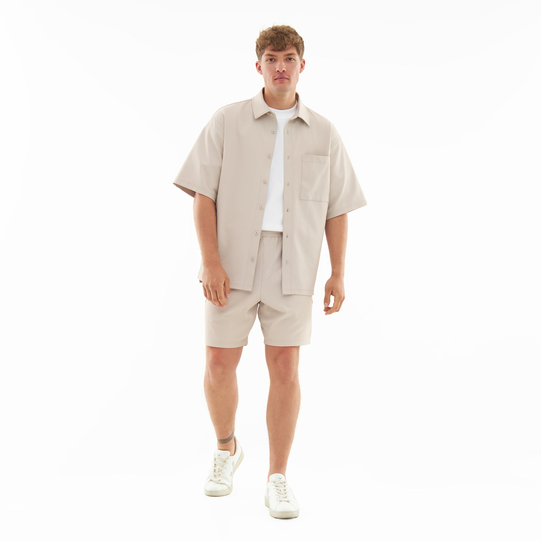 Männliches Modell trägt sandfarbenes Hemd 'Mason' mit kurzen Ärmeln und umgeschlagener Knopfleiste, kombiniert mit passenden Shorts und weißen Sneakern
