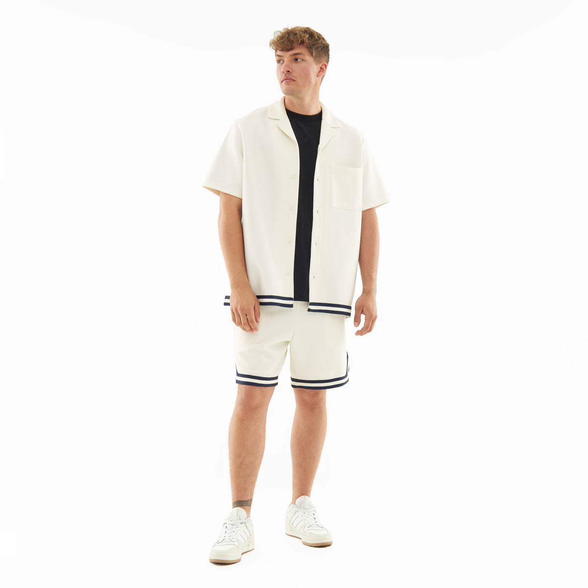 Mann trägt cremefarbenes Set 'Parker' mit kurzärmeligem Hemd und Shorts mit dunklen Streifen, weiße Sneakers