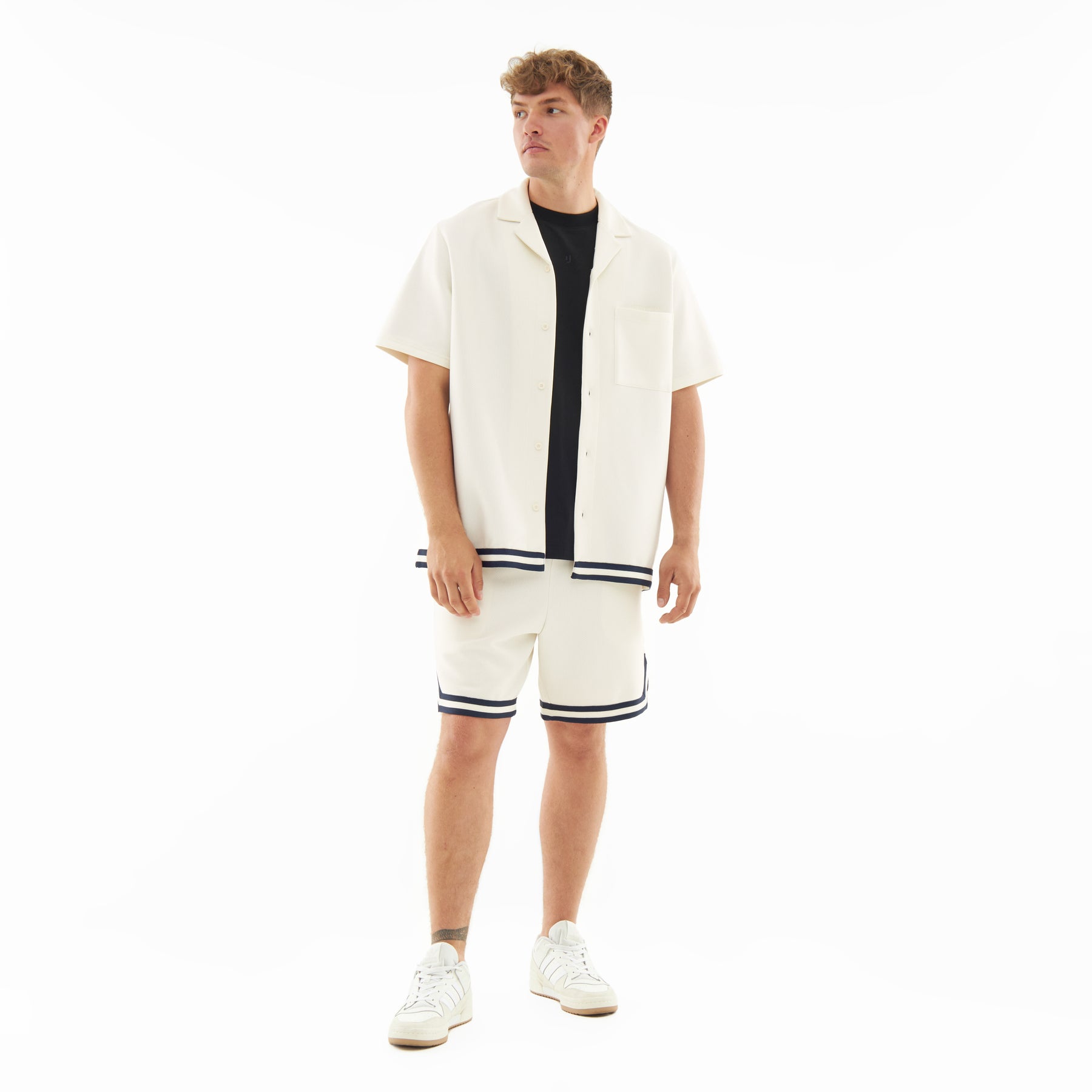Mann trägt cremefarbenes Set 'Parker' mit kurzärmeligem Hemd und Shorts mit dunklen Streifen, weiße Sneakers