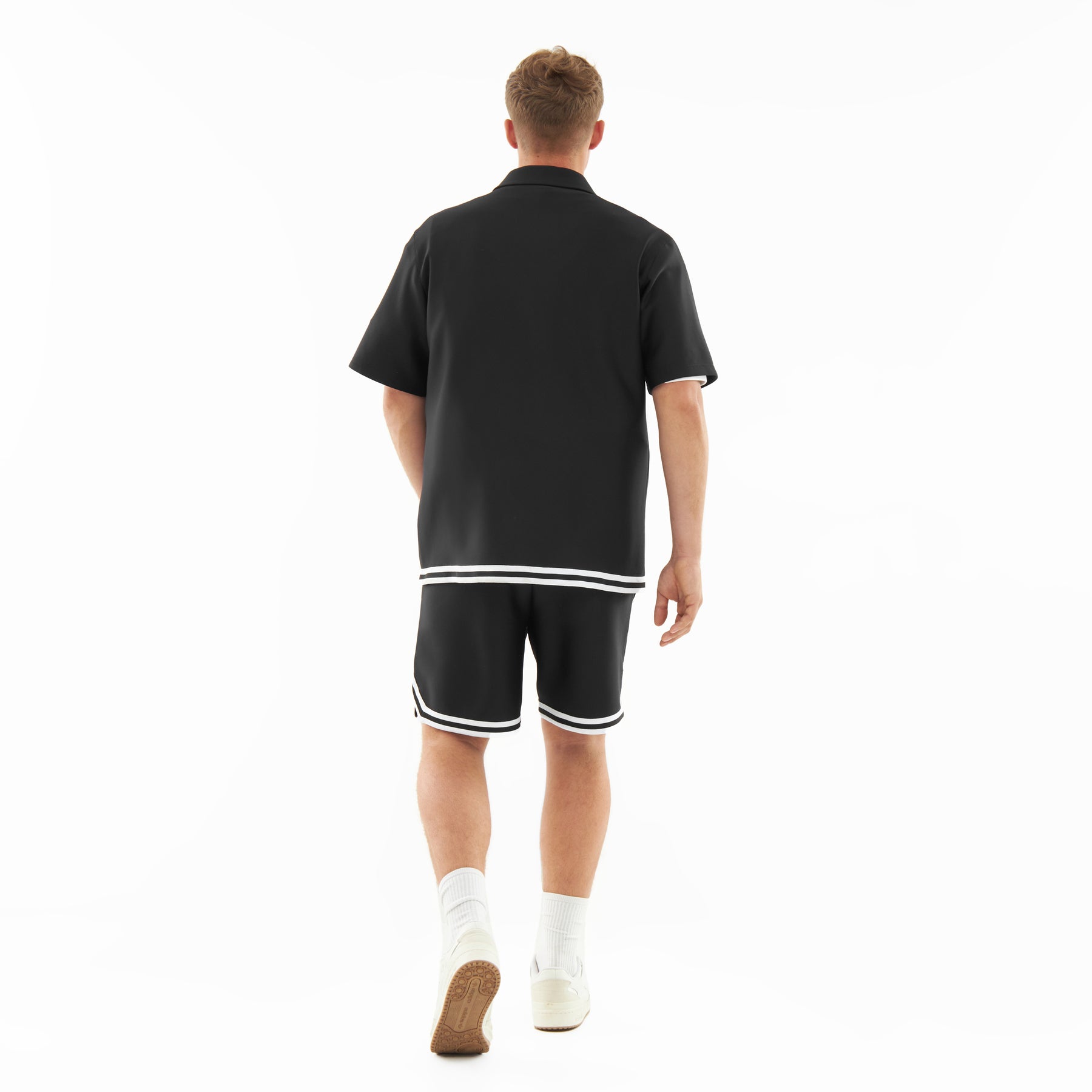 Mann von hinten in lockerem schwarzem Hemd und Shorts mit weißen Streifen am Saum, weiße Socken und Schuhe