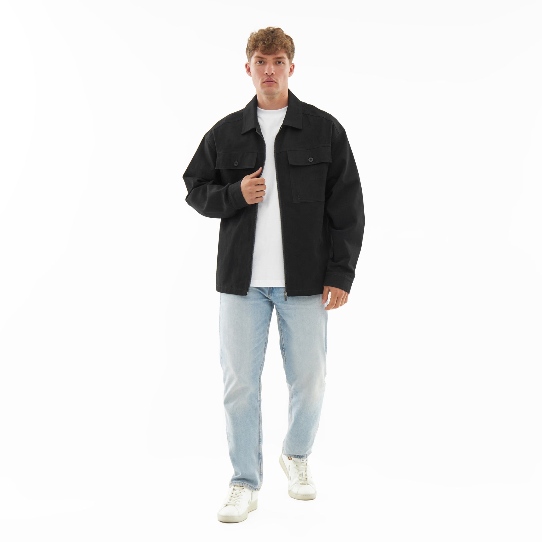 Mann trägt schwarze Baumwoll-Overshirt 'Cameron', weiße T-Shirt, helle Jeans und weiße Sneaker
