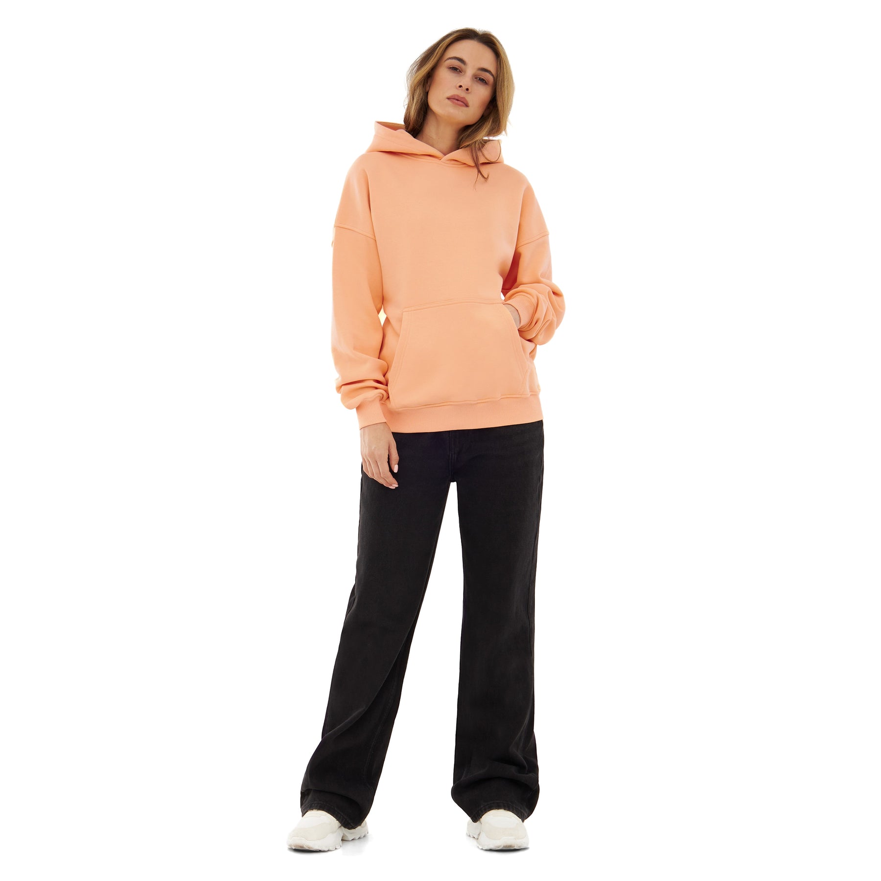 Frau trägt oversized Hoodie 'Cody' in Pfirsich mit Kapuze und Kängurutasche, kombiniert mit schwarzen weiten Hosen und weißen Schuhen