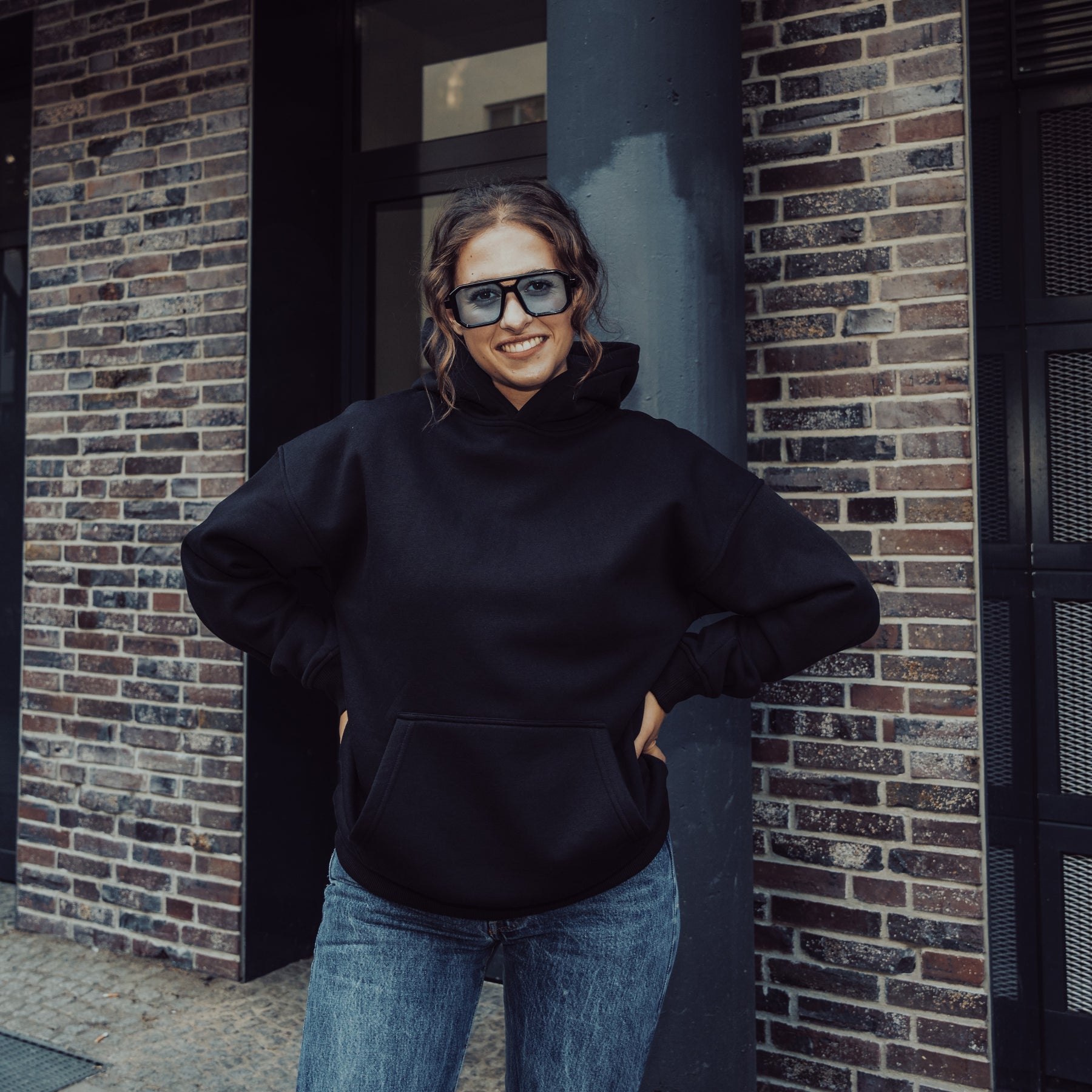 Frau trägt schwarzen Oversized Hoodie 'Cody' von Johnny Urban und blaue Jeans, steht vor Backsteinwand
