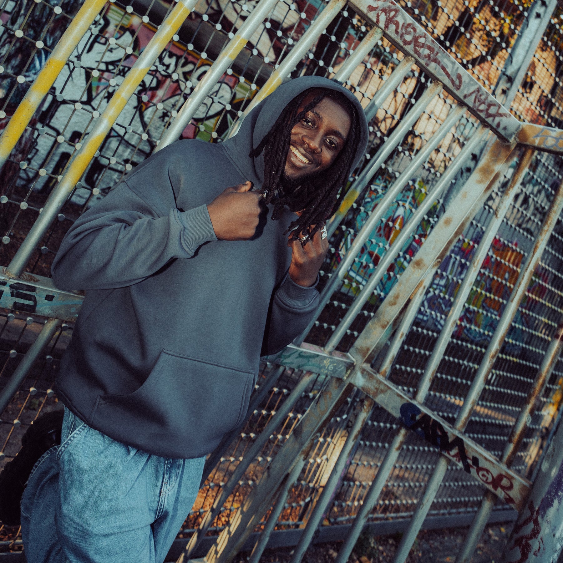 Mann trägt eine dunkelgraue oversized Hoodie 'Cody' von Johnny Urban vor einem mit Graffiti besprühten Metallzaun