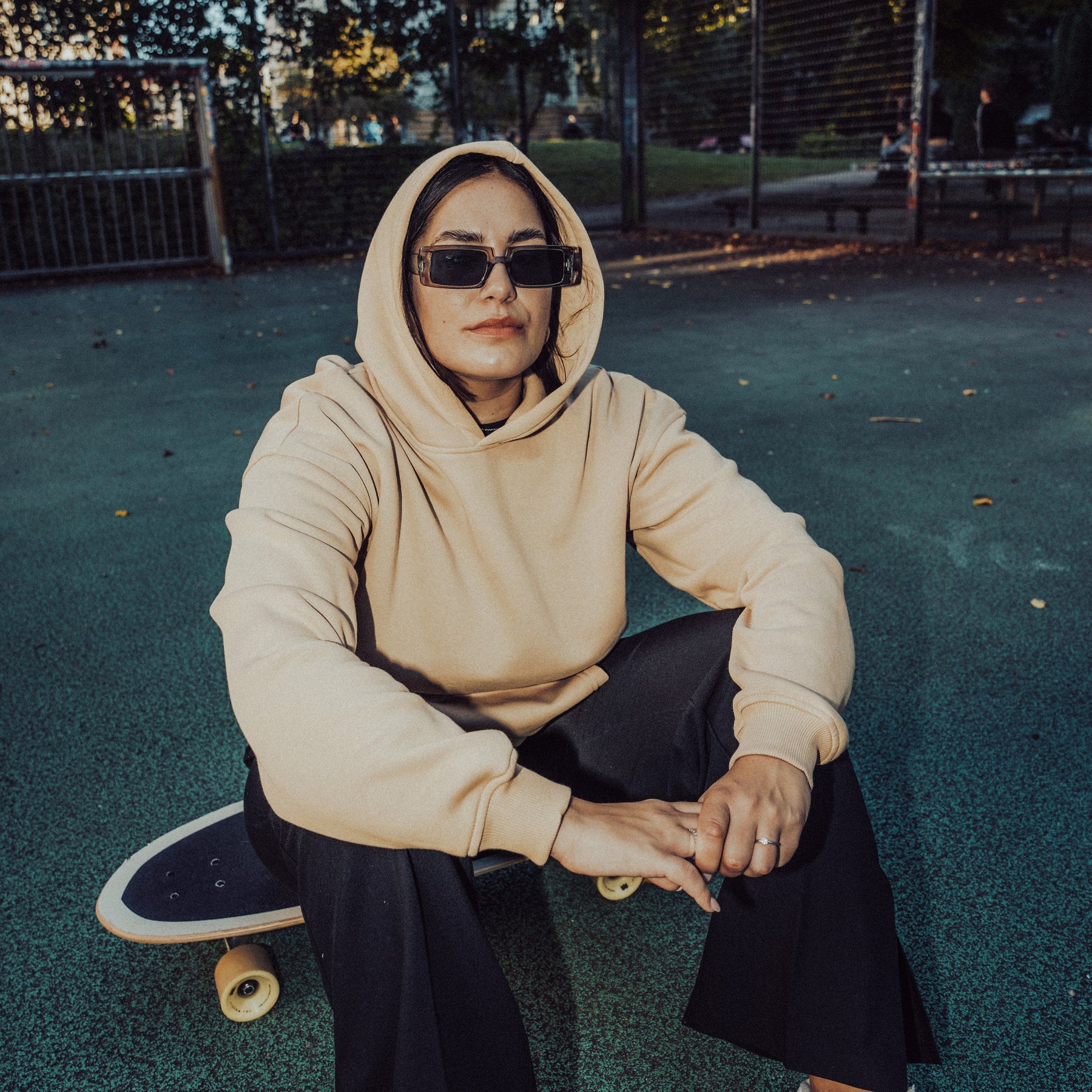 Frau trägt beigen Oversized-Hoodie 'Cody' und schwarze Sonnenbrille, sitzt auf Skateboard auf blauem Boden im Freien
