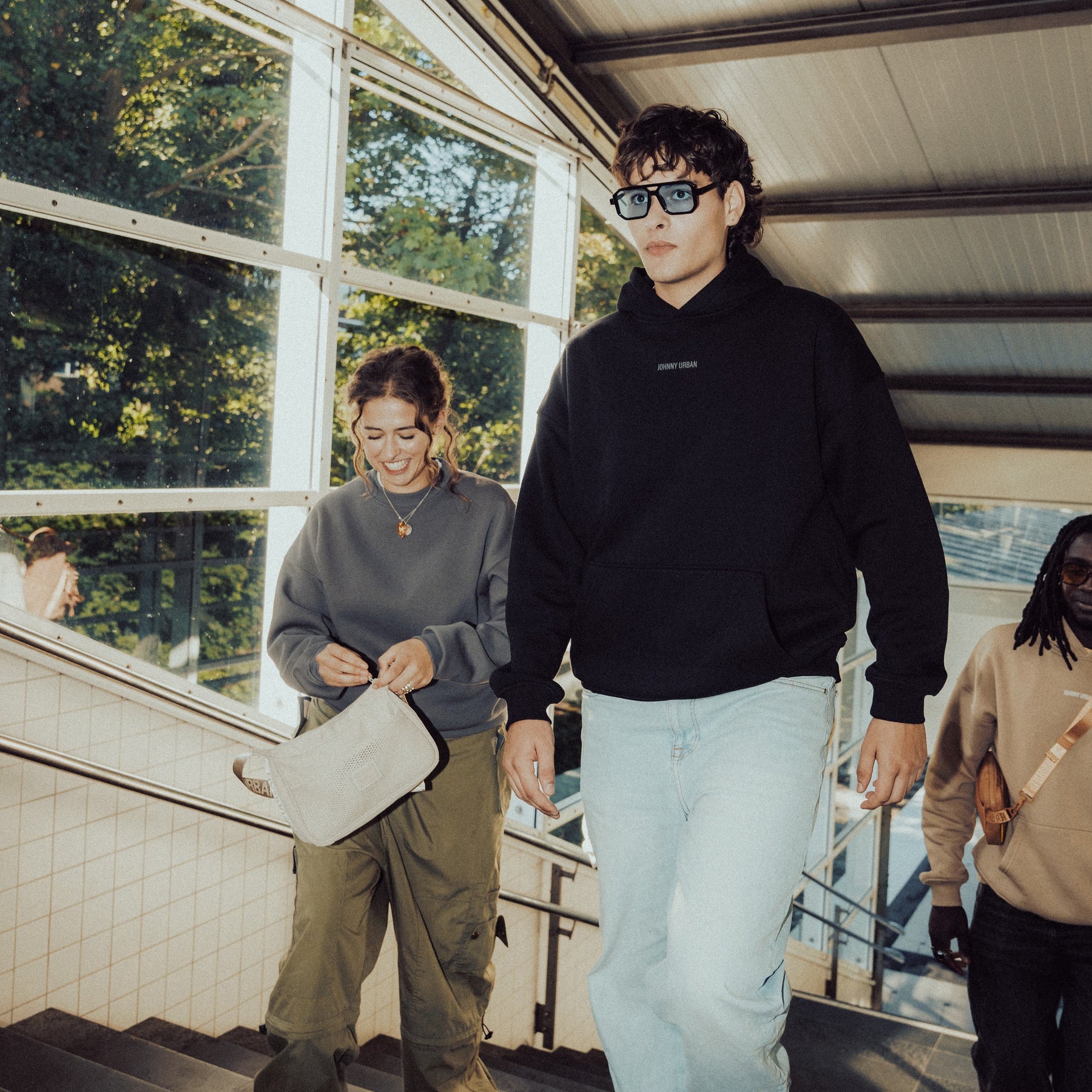 Junger Mann trägt schwarzen oversized Hoodie 'Cody' mit 'Johnny Urban' Print und helle Jeans, begleitet von zwei Personen auf Treppe