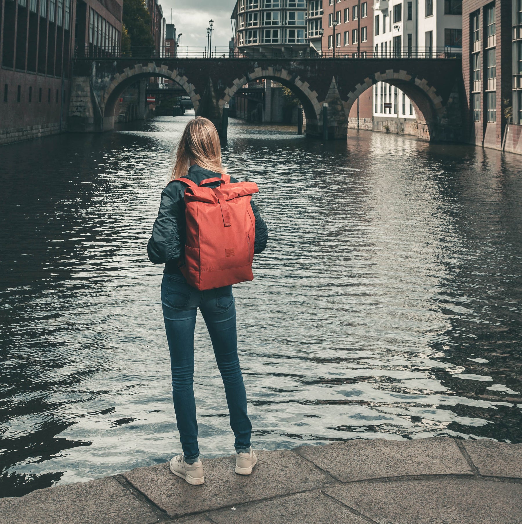 Frau mit dunkelgrüner Jacke und blauen Jeans trägt roten Johnny Urban Rolltop Rucksack 'Robin Large' an einem städtischen Flussufer