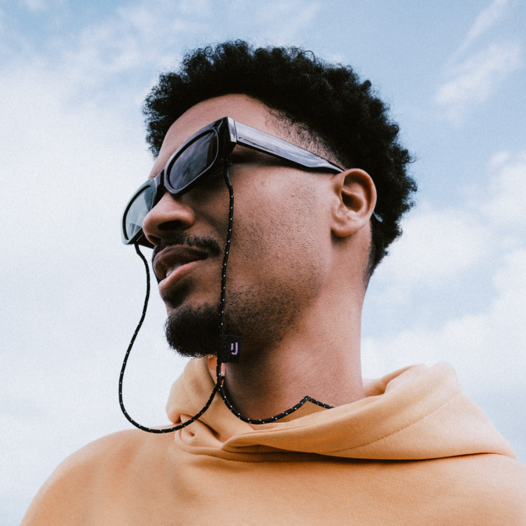 Mann mit schwarzen Sonnenbrillen mit dem Sonnenbrillenband 'Jade' in Schwarz und kleiner Logo-Anhänger am Ende