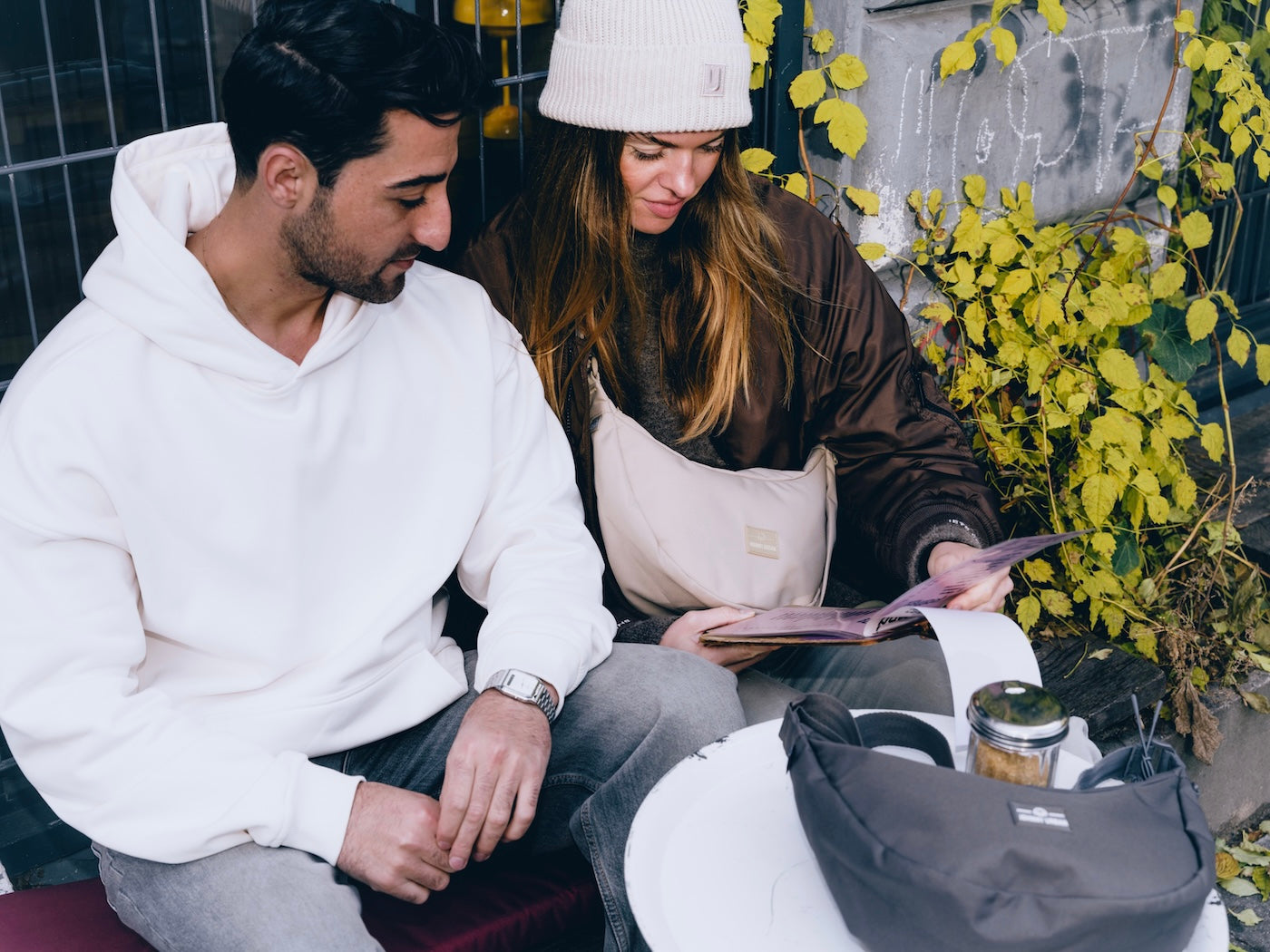 Frau trägt Half Moon Bag 'Ally Medium' in Beige, sitzt mit Mann an Tisch im Freien, sie liest Magazin