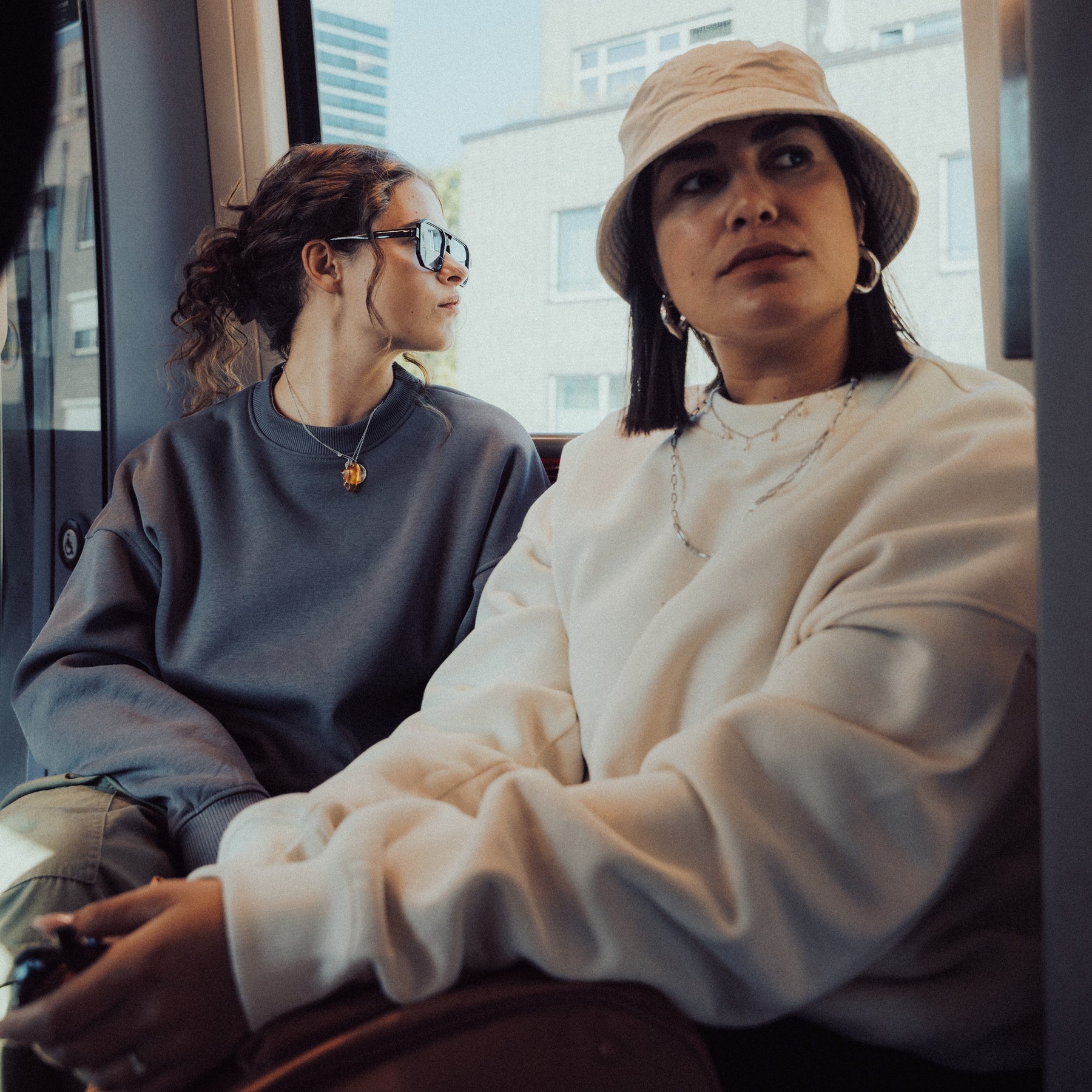 Zwei Frauen sitzen in der Straßenbahn, eine trägt ein graues Oversized Crewneck 'Carter', die andere einen weißen Pullover und einen Hut