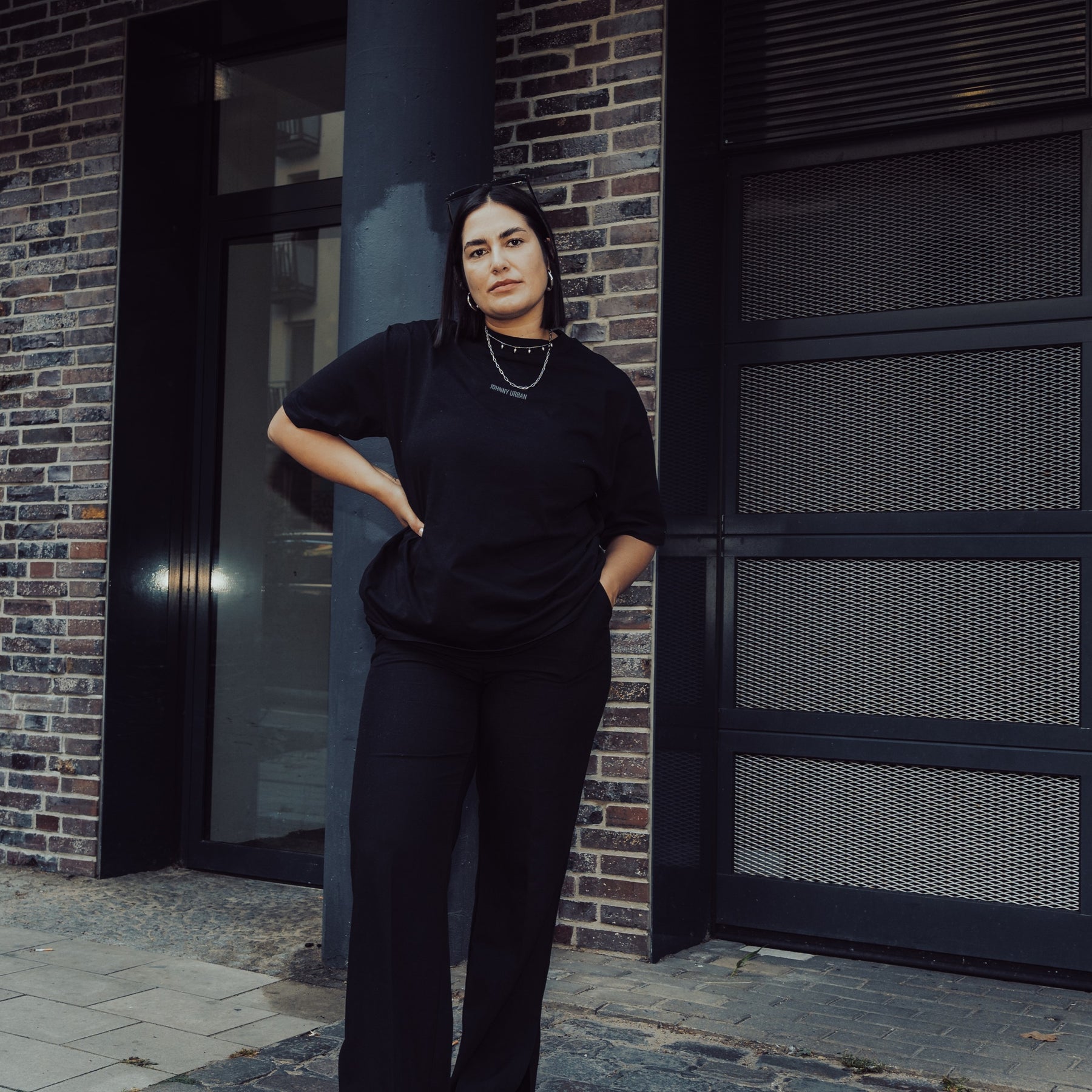 Frau trägt schwarz oversized T-Shirt 'Sammy Johnny Urban' und schwarze Hose vor urbanem Hintergrund