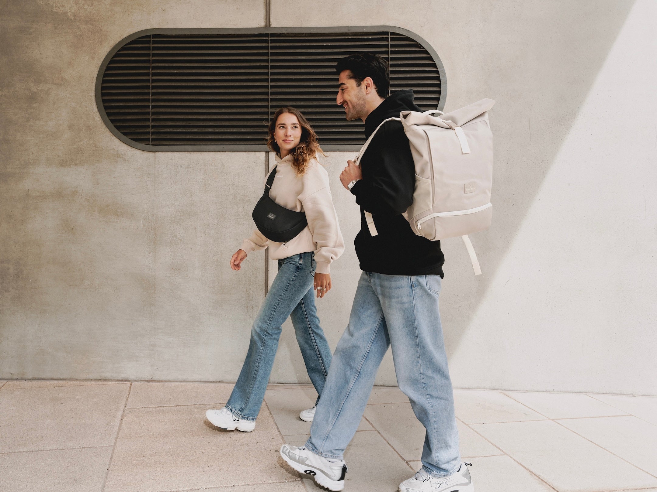 Frau mit Half Moon Bag "Alley Medium" und Mann mit Rolltop Rucksack "Allen Large" gehen und unterhalten sich.