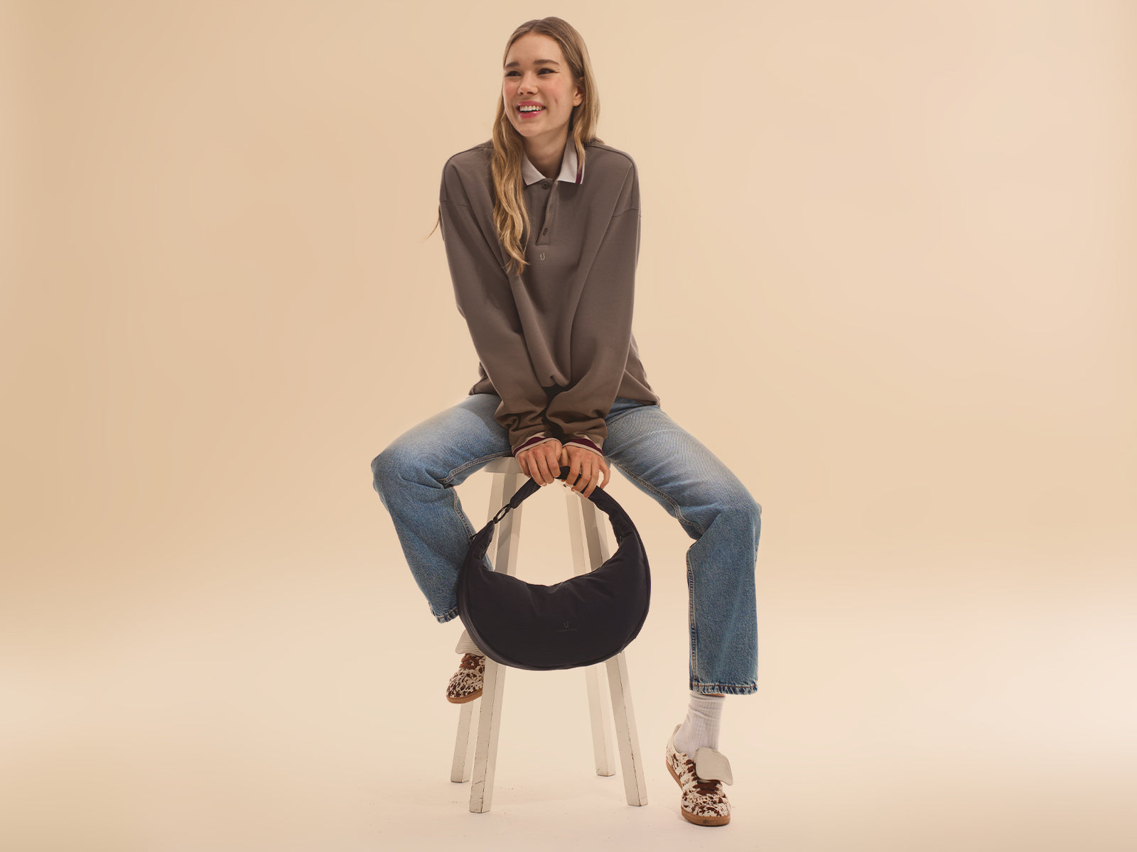Frau sitze auf einem Hocker, lächelt und hält eine schwarze Half Moon Bag "Lou Large" in den Händen.
