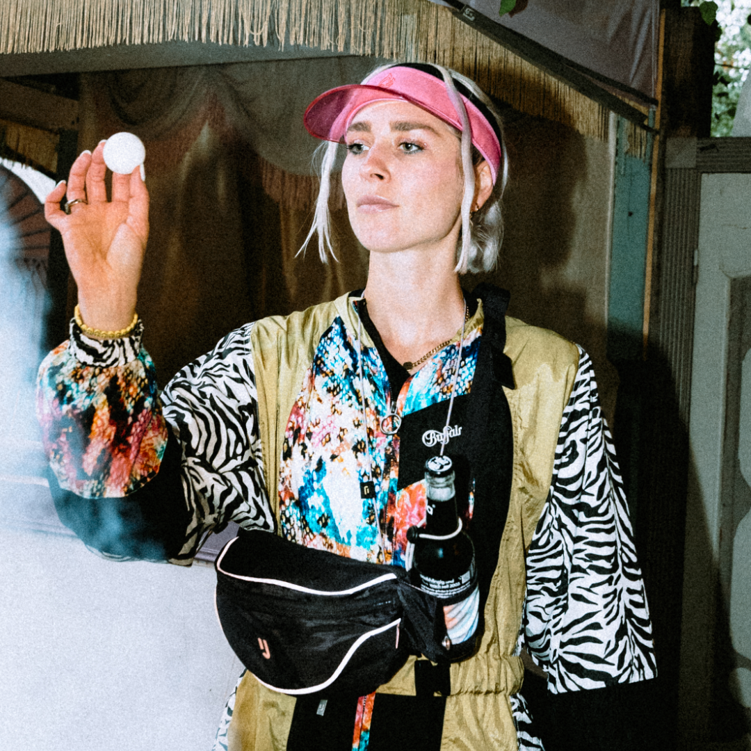Frau trägt pinke Sun Visor 'Joy' und bunte Jacke mit Zebra-, Blumenmuster, dazu schwarze Bauchtasche und Bierflasche.