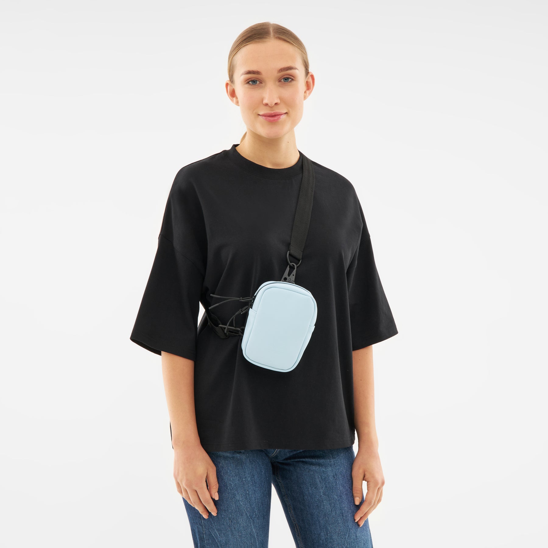 Frau trägt hellblaue Front Pack 'Toby' Crossbody-Tasche über schwarzem T-Shirt und blauen Jeans