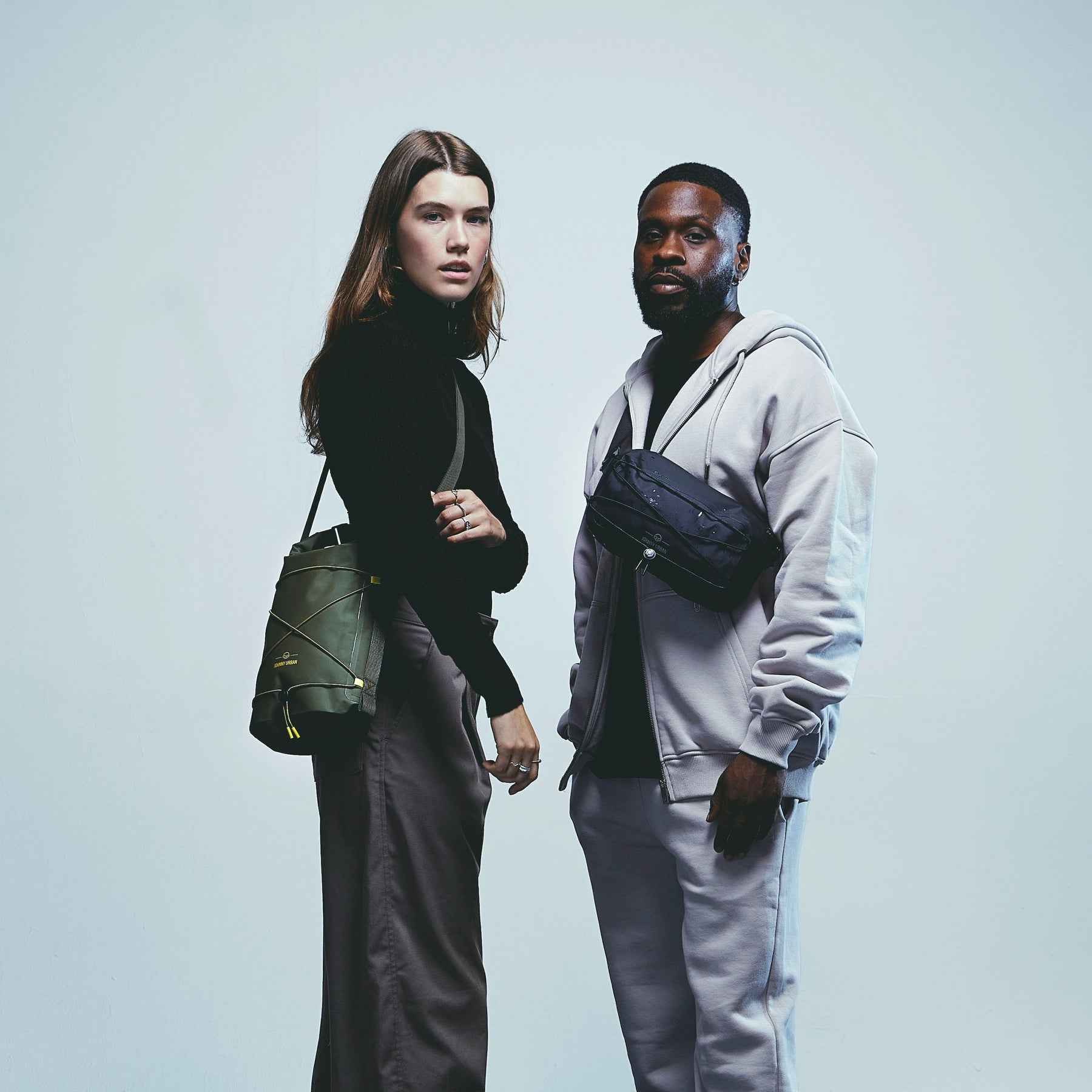 Frau mit schwarzer Lenkertasche 'Arlo' in Olivgrün über der Schulter, Mann mit schwarzer Umhängetasche 'Arlo' in Kapuzenjacke und Jogginghose